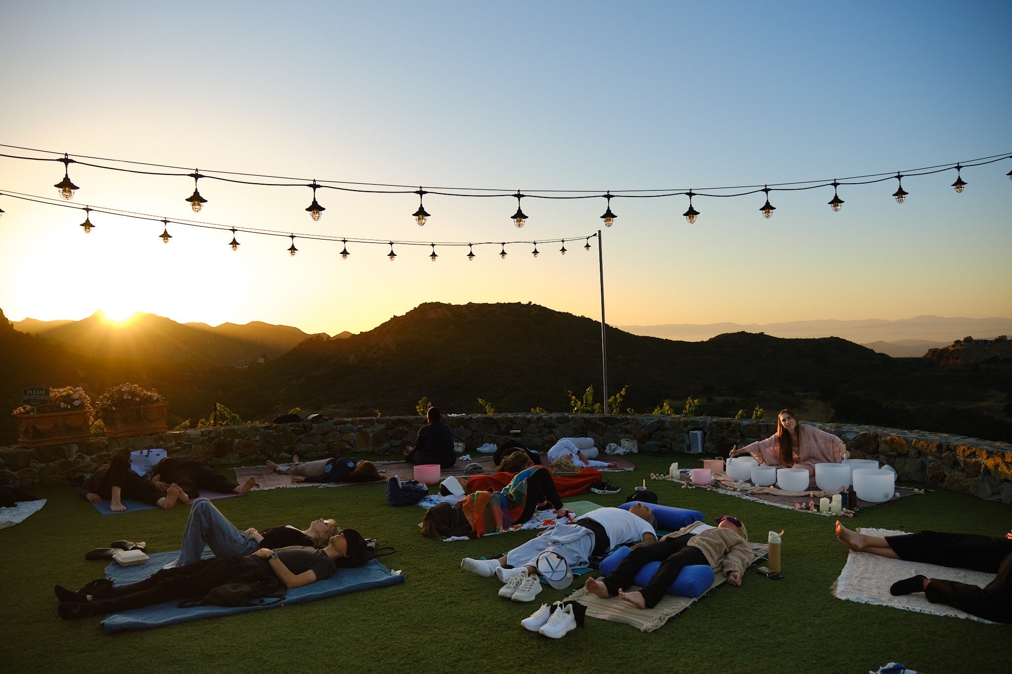 People practicing yoga outdoors during sunset on a grassy hill, with string lights hanging overhead and mountain scenery in the background.