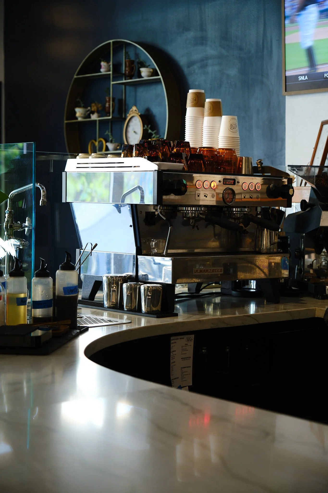 A coffee shop counter with a coffee machine, stacks of paper cups, and various bottles and utensils. A dark blue wall with a decorative shelf holding teapots and cups, and a television screen showing a baseball game.