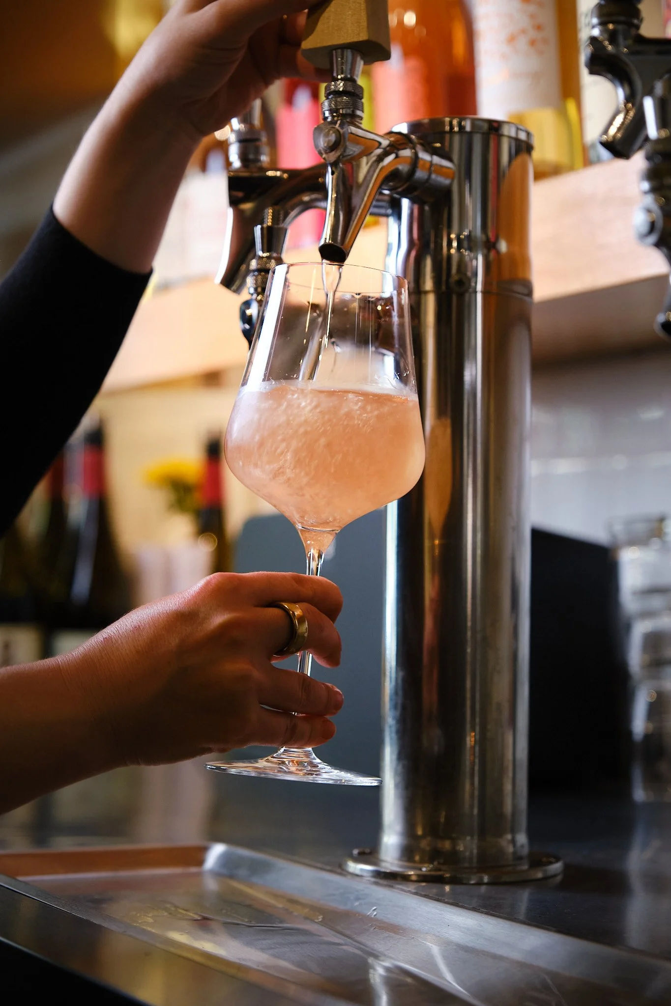 A person holding a wine glass under a beer tap, filling it with a pinkish, fizzy beverage in a bar or restaurant setting.