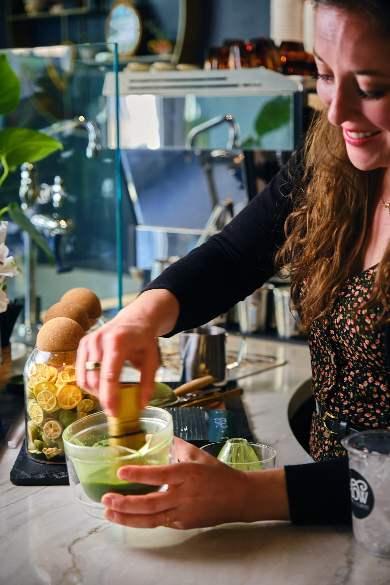A woman pouring a drink with a green garnish, with fresh citrus slices and herbs in glass containers nearby, in a modern kitchen or bar setting.