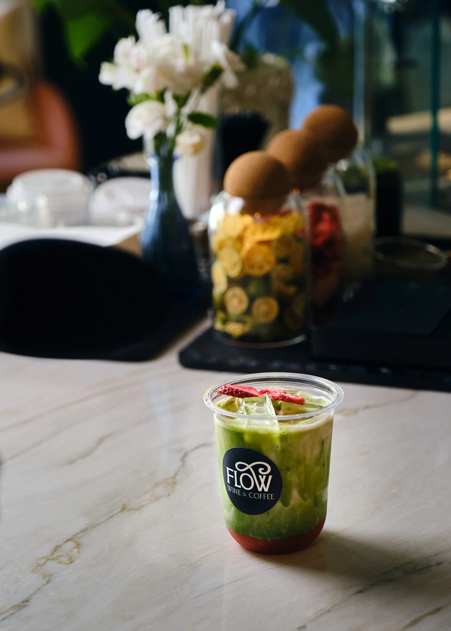 A colorful layered drink with a straw on a light marble countertop, with a blurred background of a floral arrangement and kitchen objects.