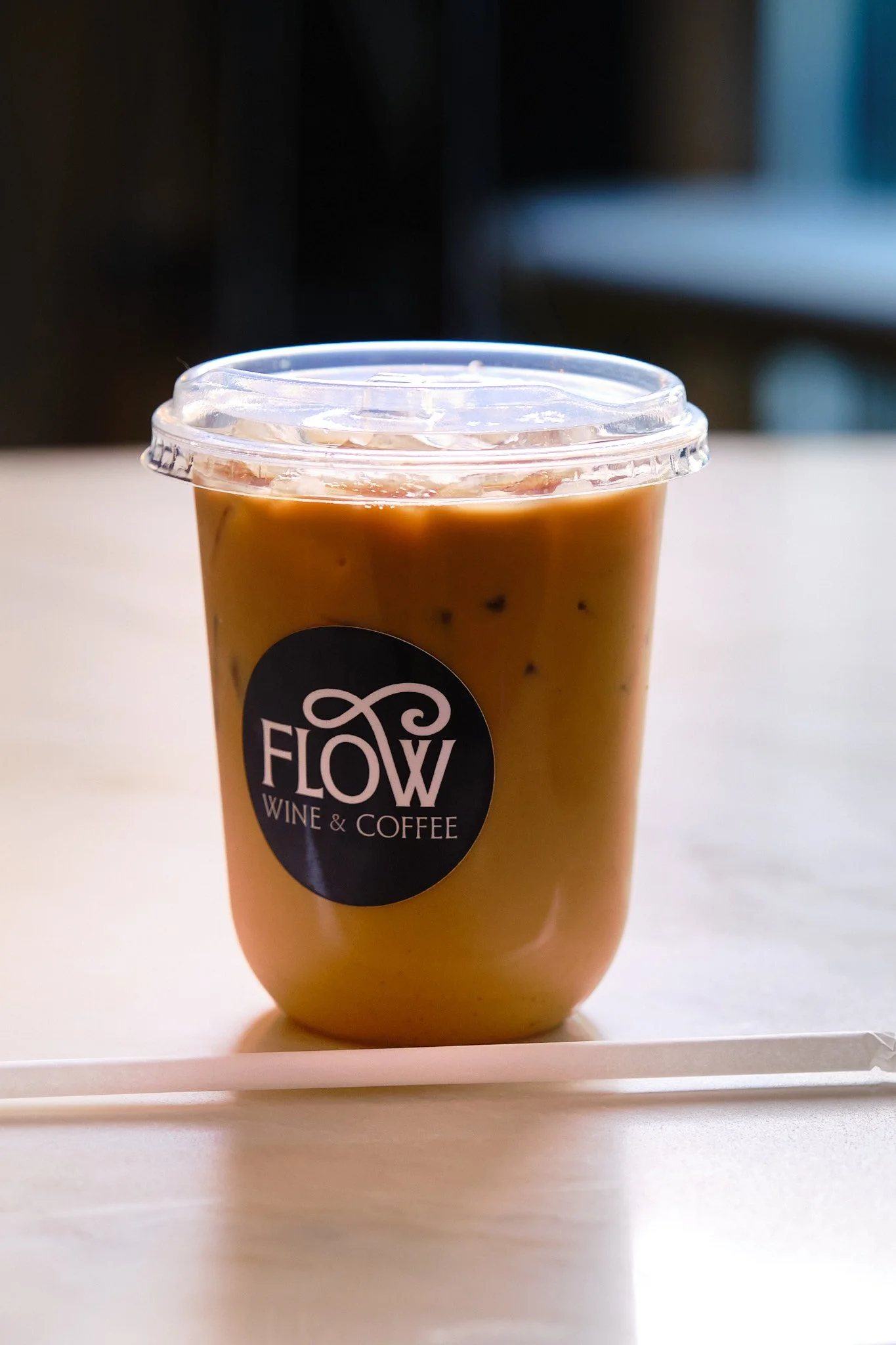 Plastic cup filled with iced coffee, featuring a black label with white text reading 'FLOW WINE & COFFEE', placed on a white surface with a blurred indoor background.