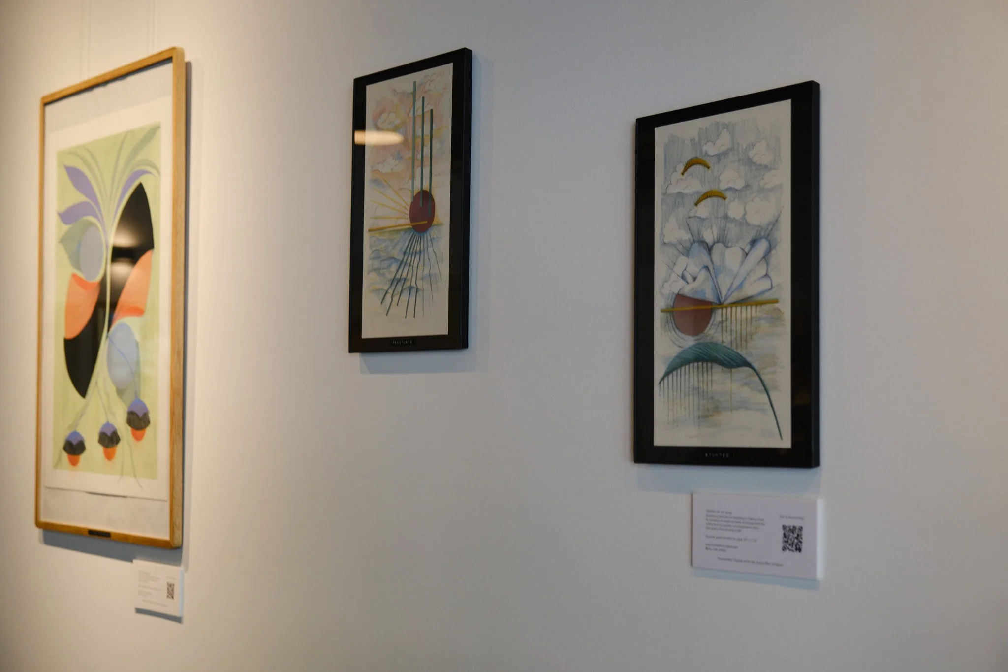 Three framed abstract art pieces hanging on a gallery wall with text and QR code labels below each.