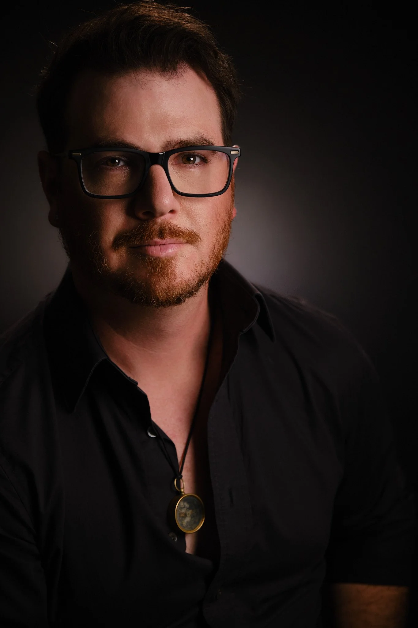 A man with brown hair, brown eyes, and a red beard wearing black glasses and a black shirt, with a dark background.