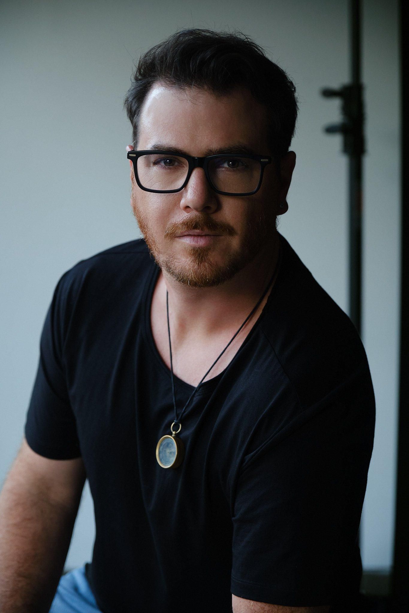 A young man with dark hair, glasses, and a beard wearing a black shirt and a locket necklace, sitting indoors with a neutral background.