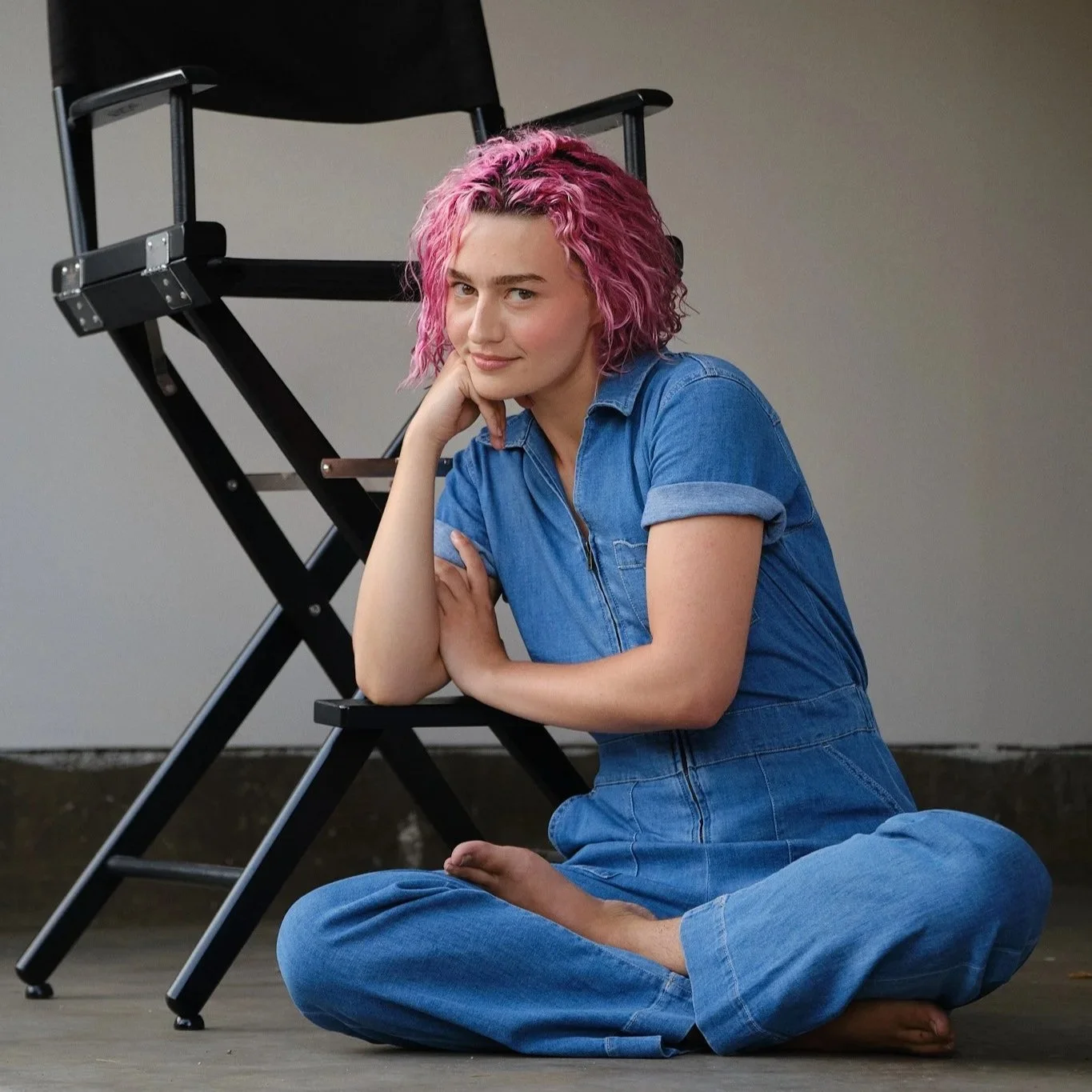 A woman with curly pink hair sitting cross-legged on the floor, resting her chin on her hand, in front of a black folding chair against a neutral background.