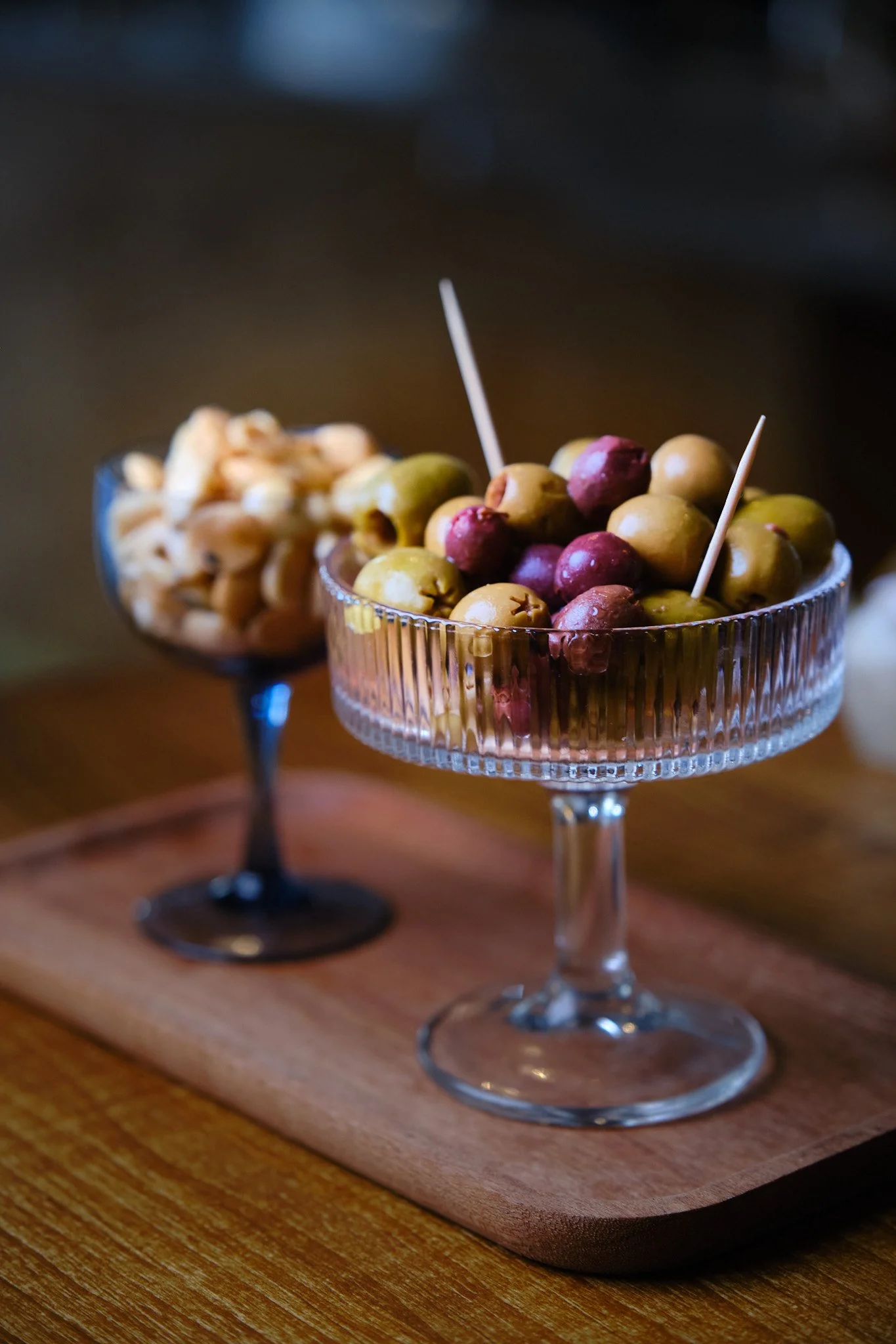 Two vintage-style glass bowls filled with green and purple olives, placed on a wooden serving board.