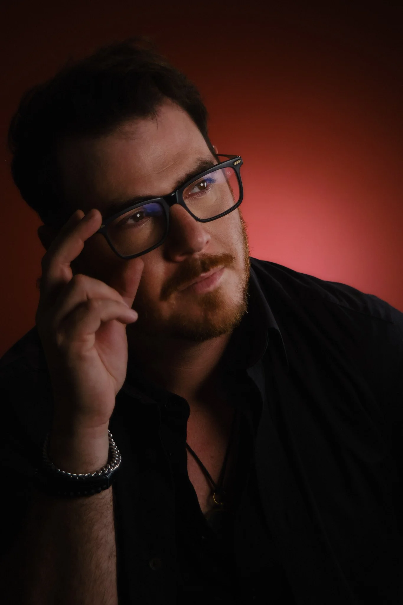 A man with brown hair, glasses, and a beard, wearing a black shirt, resting his head on his hand against a dark reddish background.