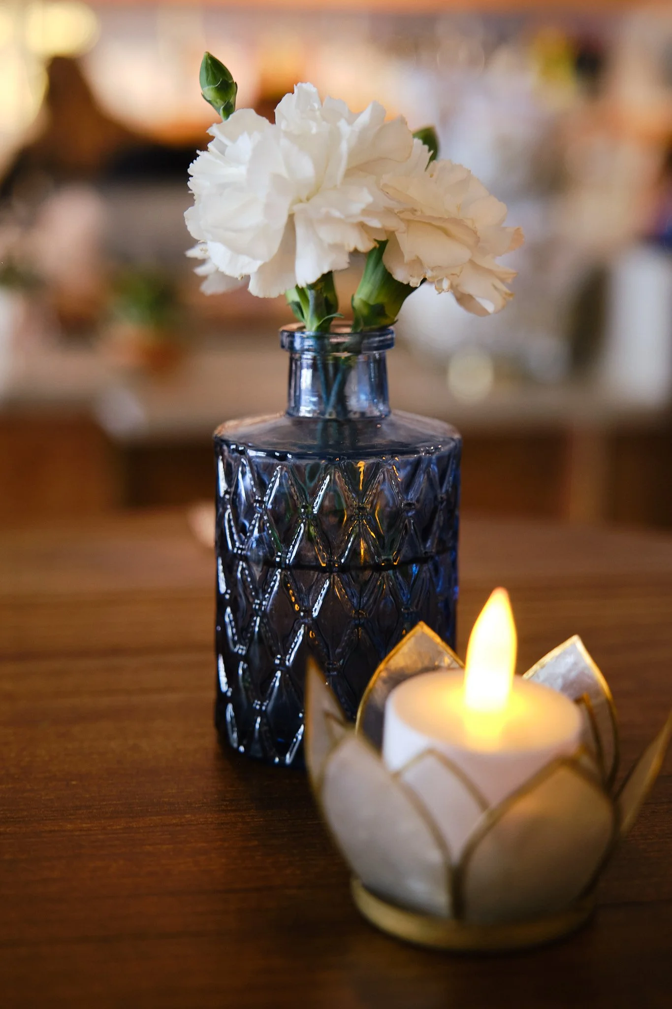 A white carnation flower in a textured blue glass bottle on a wooden surface, with a lit candle in a gold holder nearby.