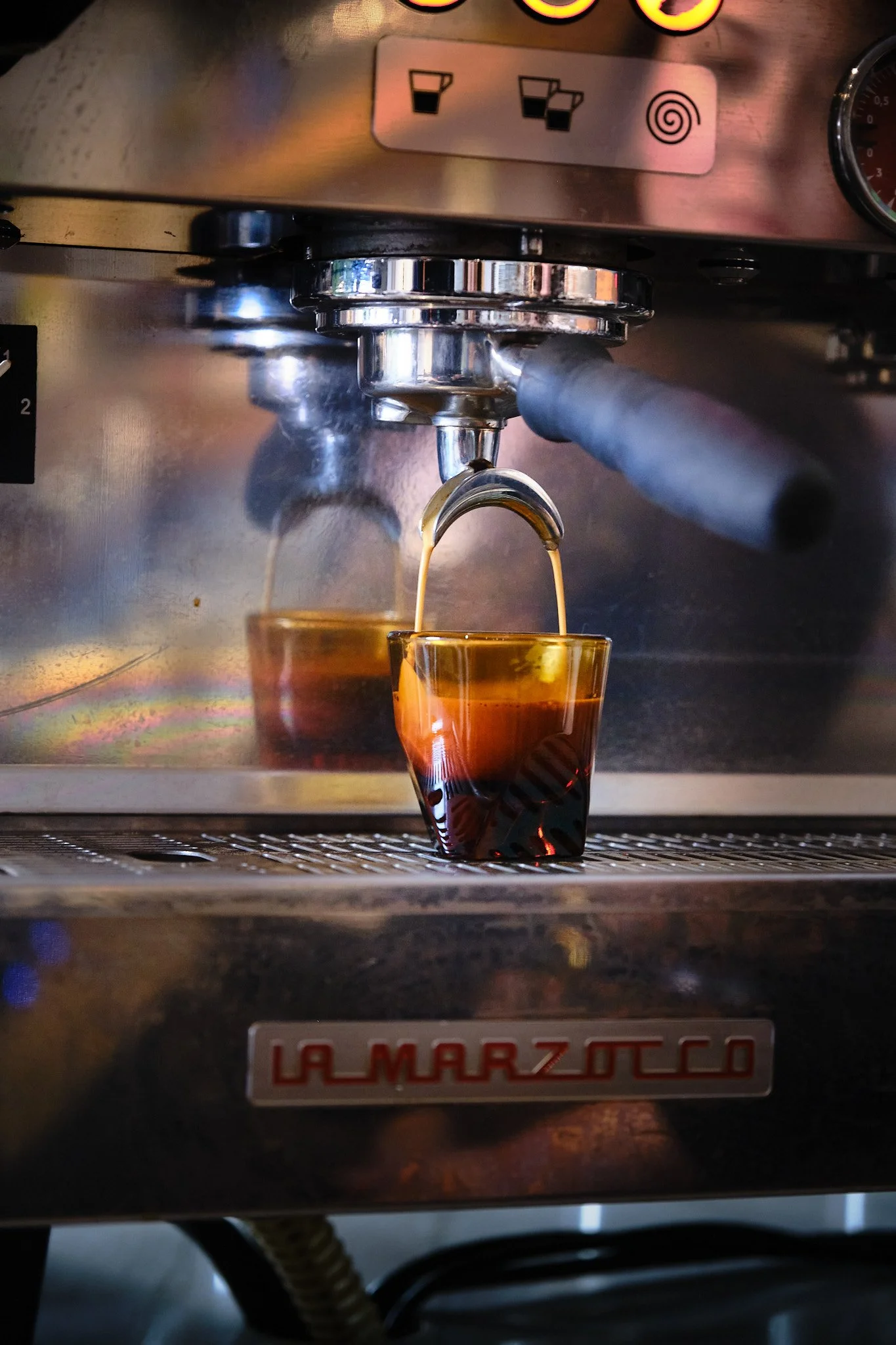 Espresso coffee is being brewed in an espresso machine, with dark coffee pouring into a glass cup on the drip tray.