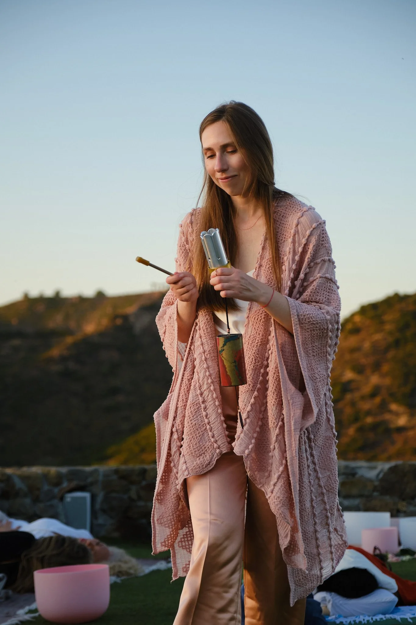A woman standing outdoors during sunset, holding a metal container and a stick, with blankets and cushions on the ground behind her.