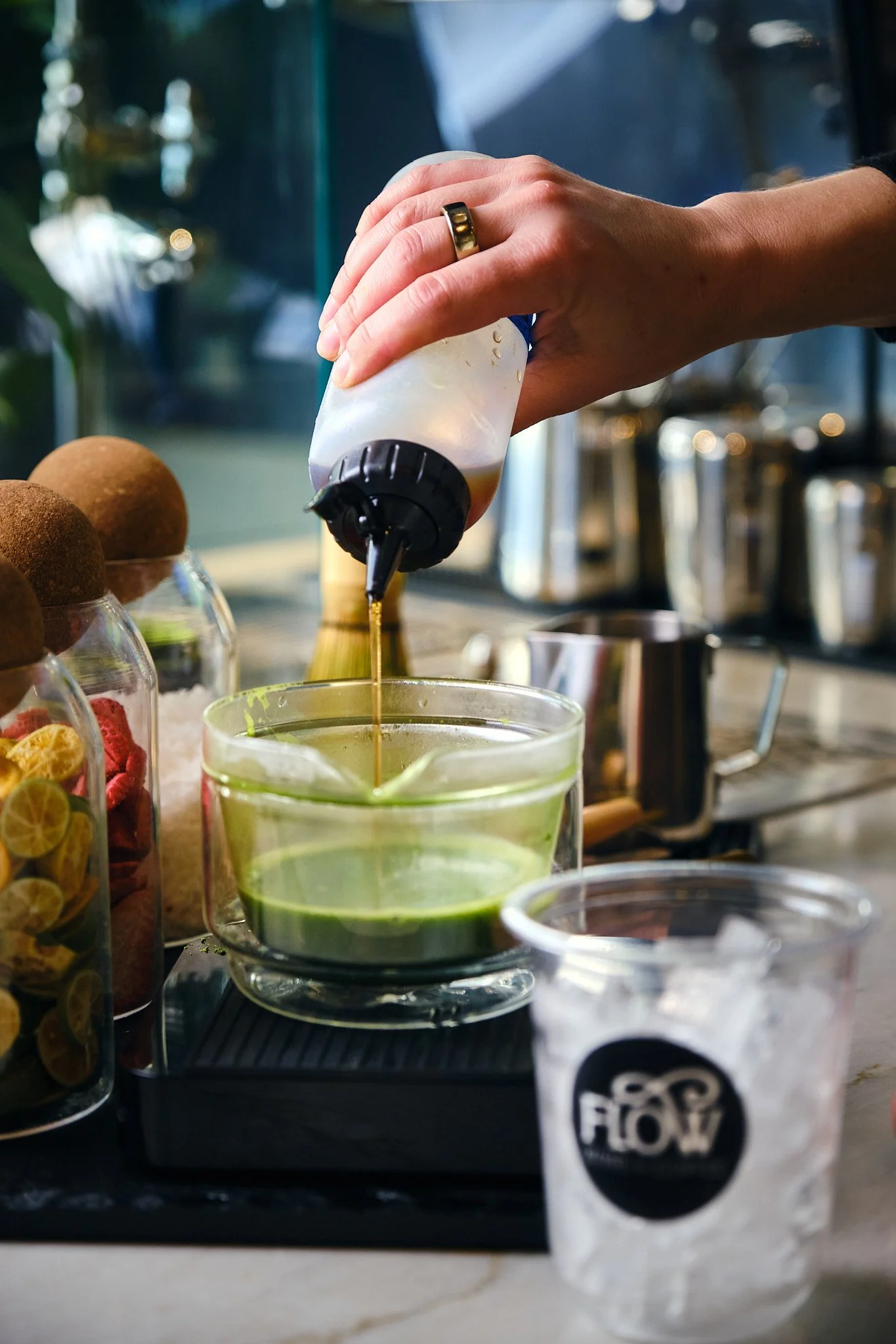 Person squeezing liquid from a squeeze bottle into a glass with green liquid. Various bottles and jars with food items, and a branded paper cup are on the counter.