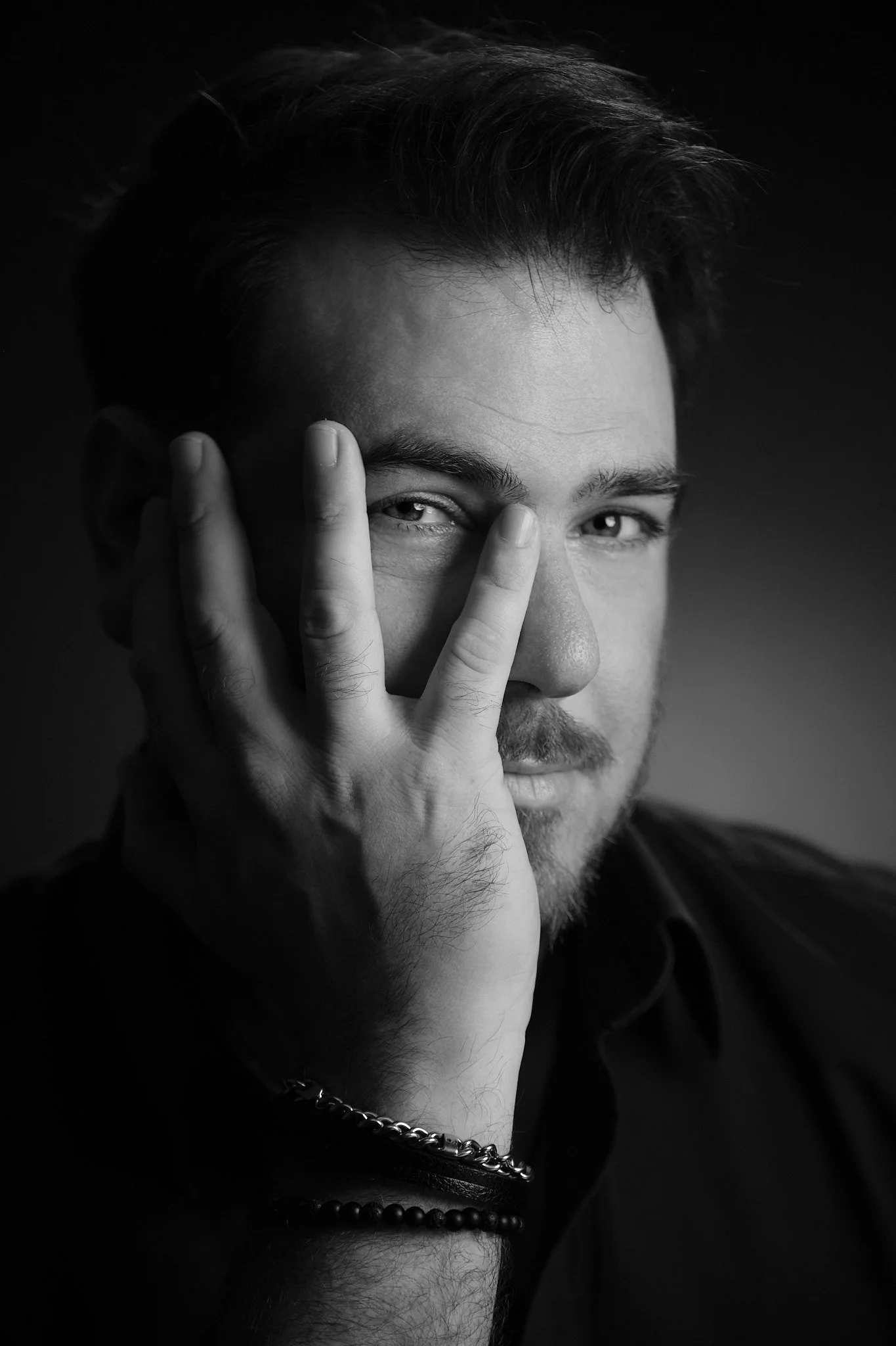 Black and white close-up portrait of a man with dark hair, a light beard, and a contemplative expression, with one hand partially covering his face, wearing jewelry including a chain and beads.