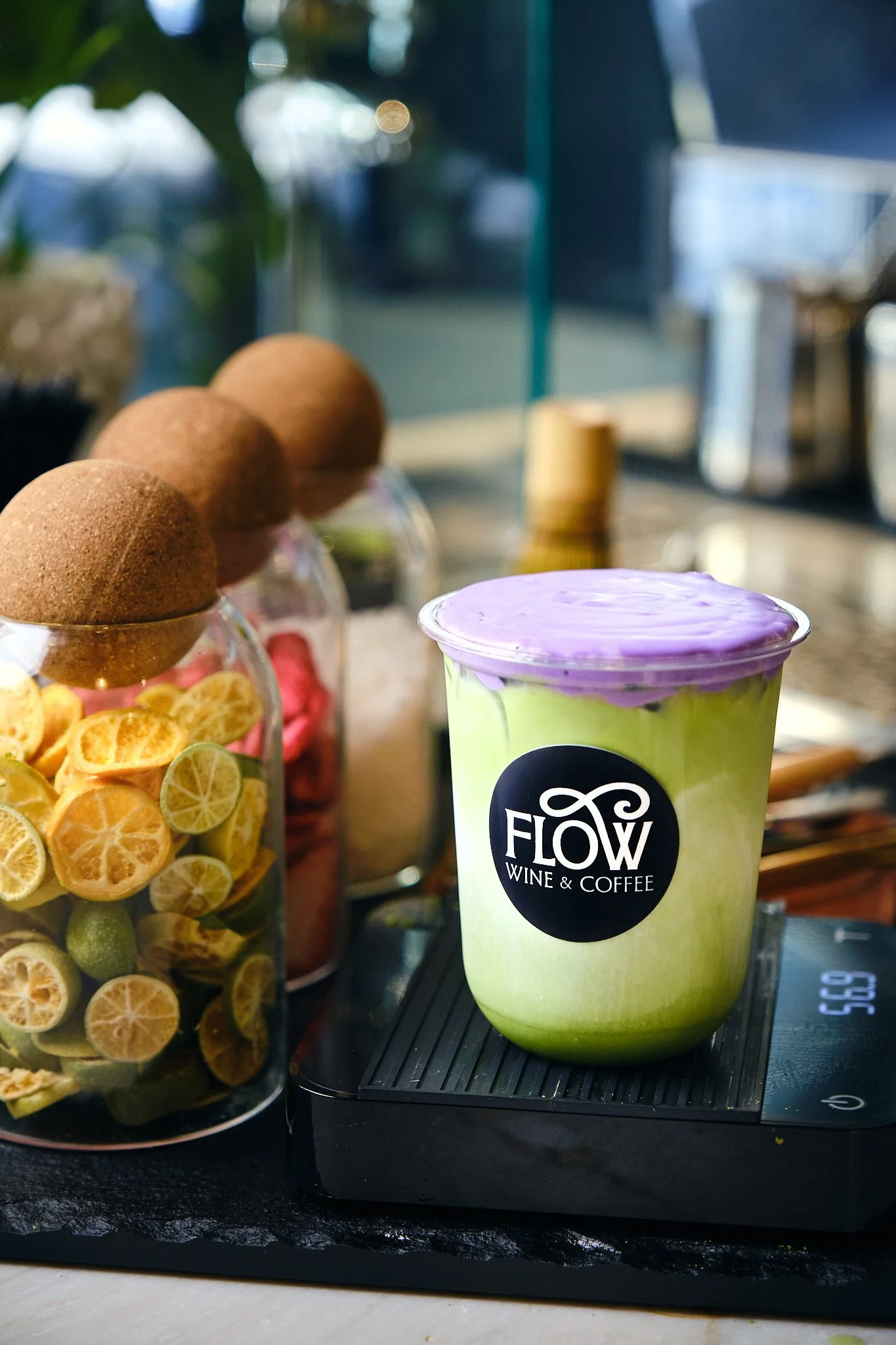 A beverage cup with 'Flow Wine & Coffee' logo on it placed on a digital scale, with a jar of dried lemon slices and a jar of brown eggs in the background.