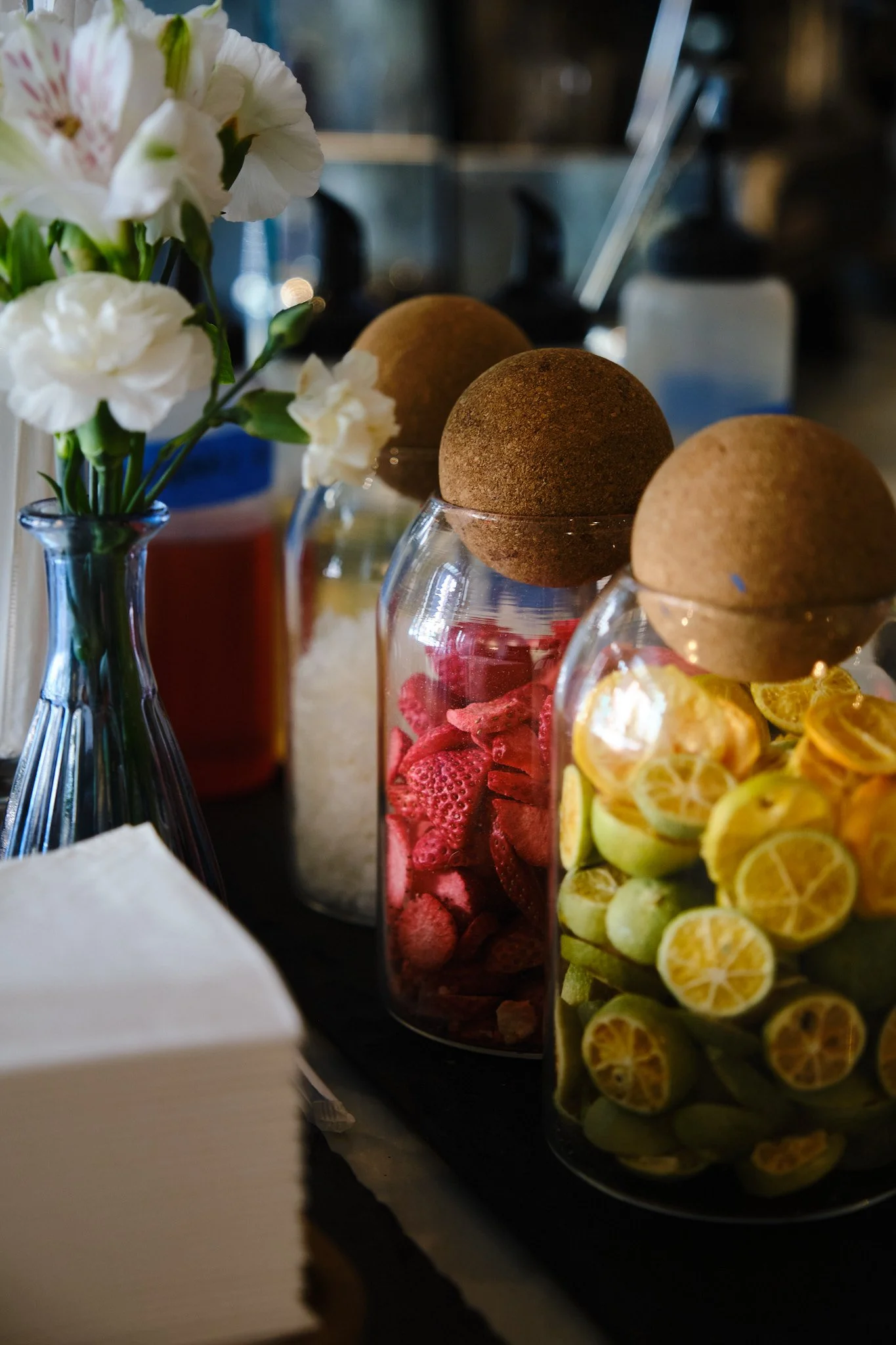 A close-up of a vase with white flowers on the left, and three glass jars filled with colorful candies and fruit slices on the right in a dark setting.