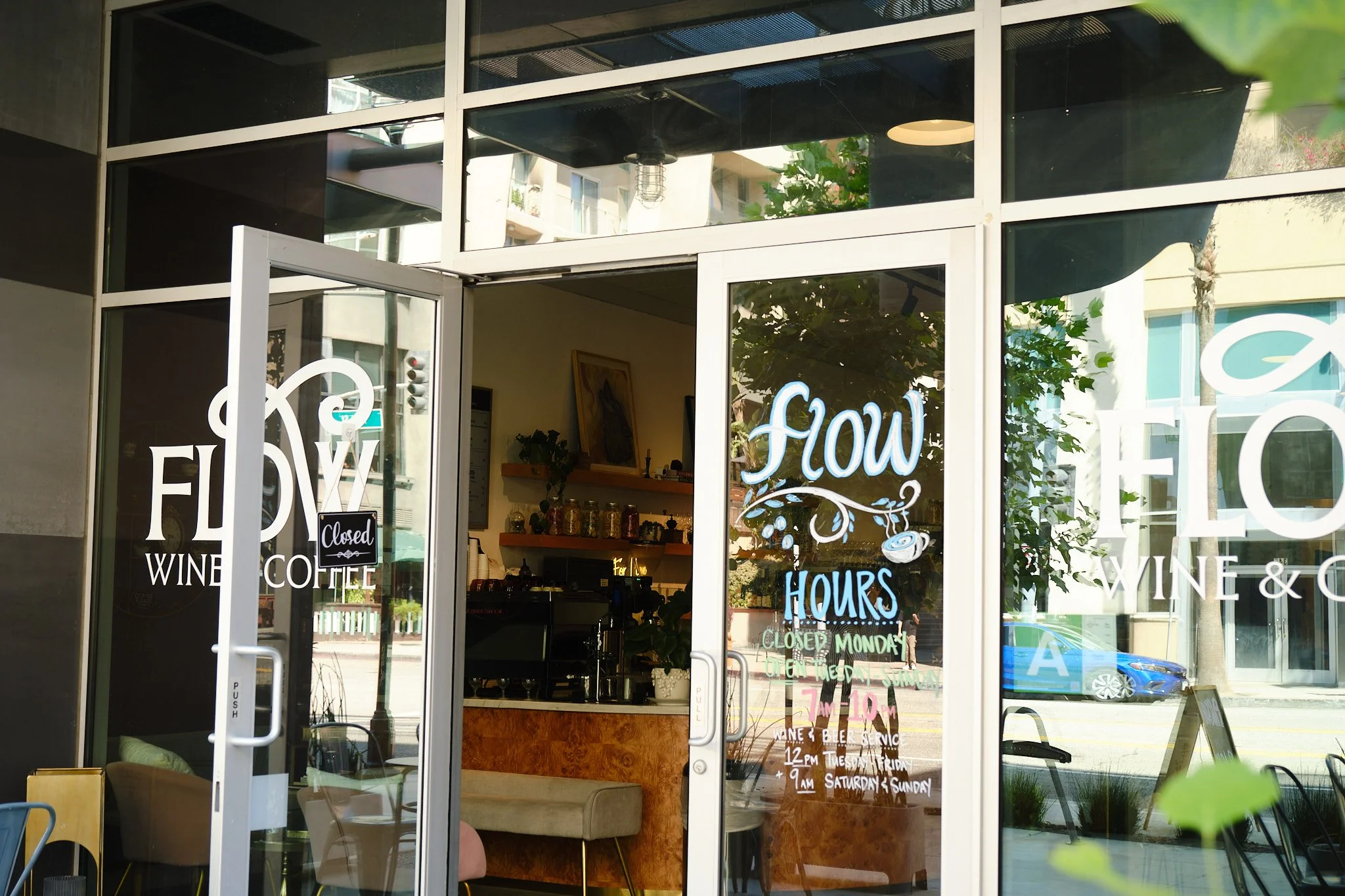 The storefront of a cafe named 'Flynn Wine + Coffee' with glass doors and windows, displaying handwritten signs about its hours of operation and closed days. Inside, there are shelves with jars, a countertop, and some greenery.