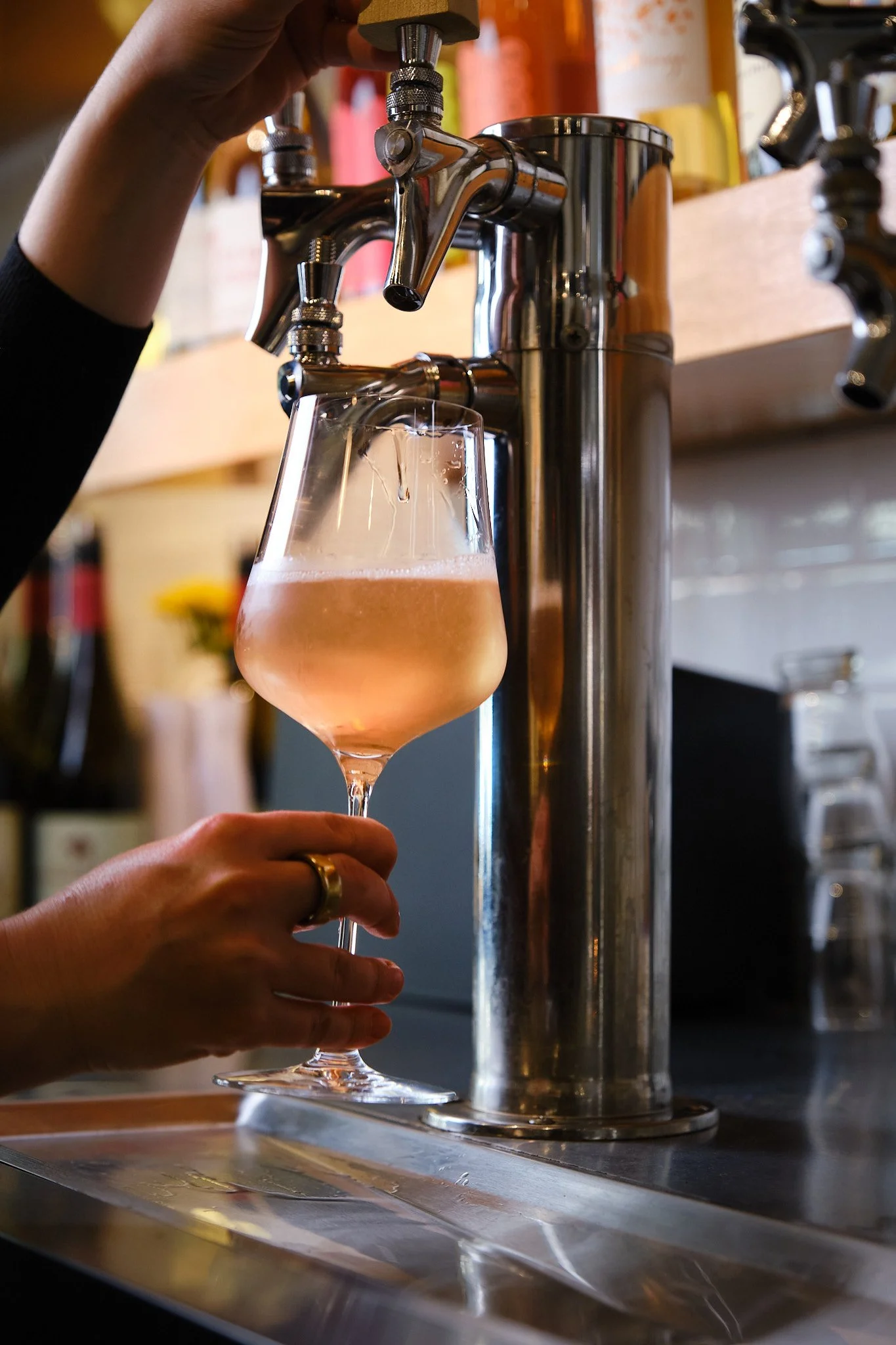 A person is filling a glass with rosé wine from a tap at a bar or restaurant with a wooden shelf in the background.