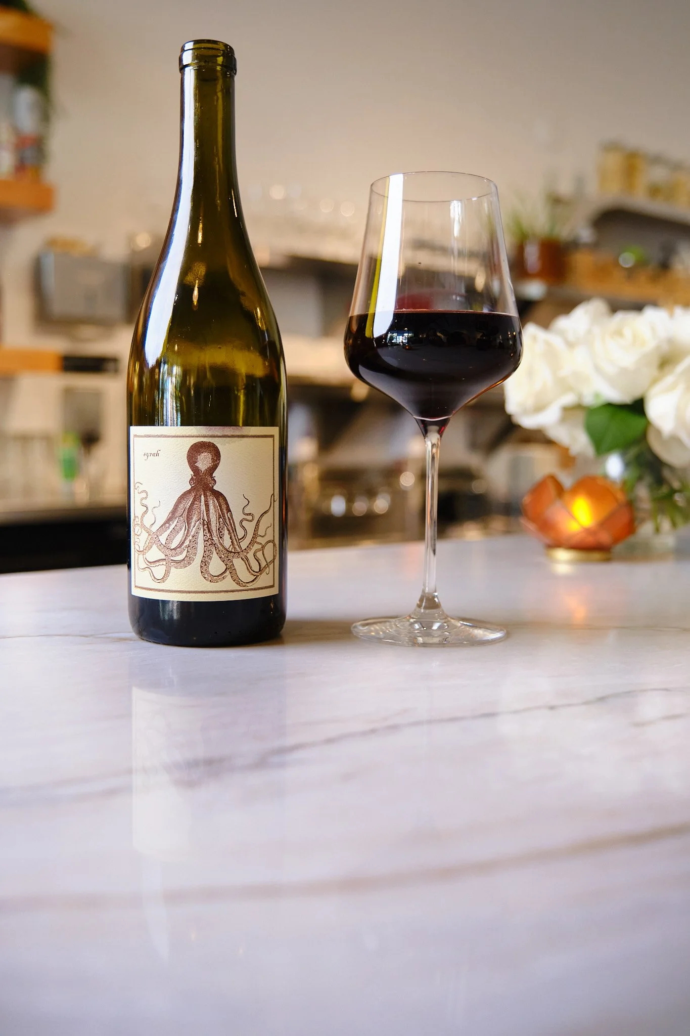 A bottle of red wine and a glass filled with red wine on a kitchen countertop, with a blurred kitchen background and a bouquet of white roses.