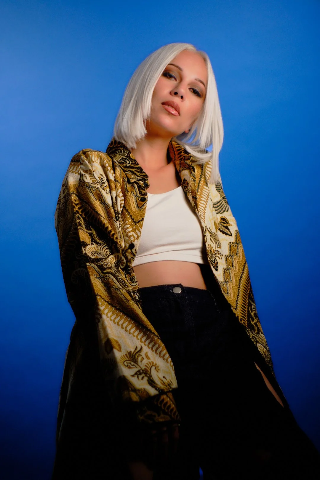 Young woman with platinum blonde hair, wearing a patterned gold and black jacket over a white crop top, posing against a blue background.