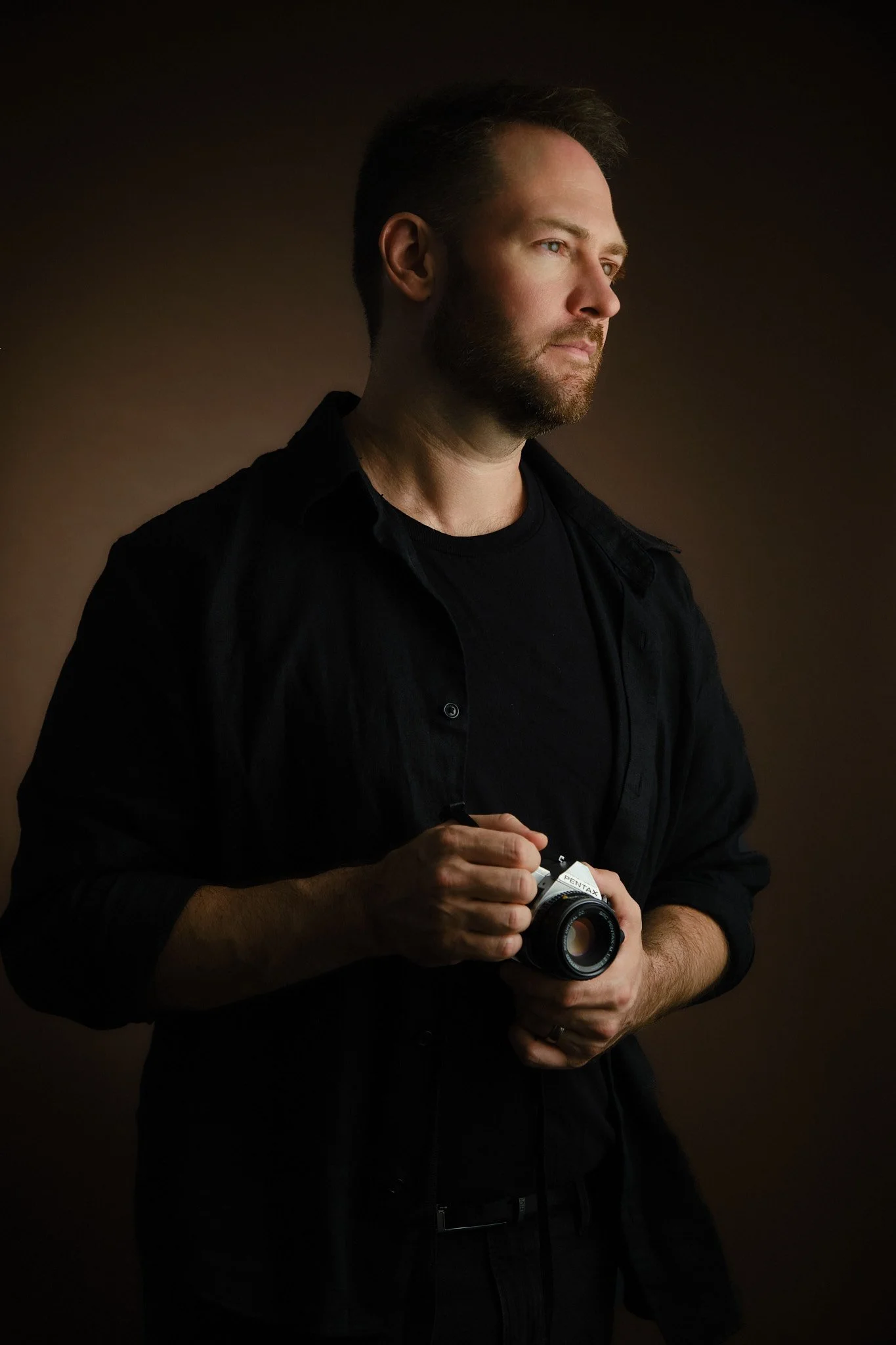 A man with a beard and short brown hair looking to the right, holding a vintage camera with both hands, wearing a black shirt and standing against a dark background.
