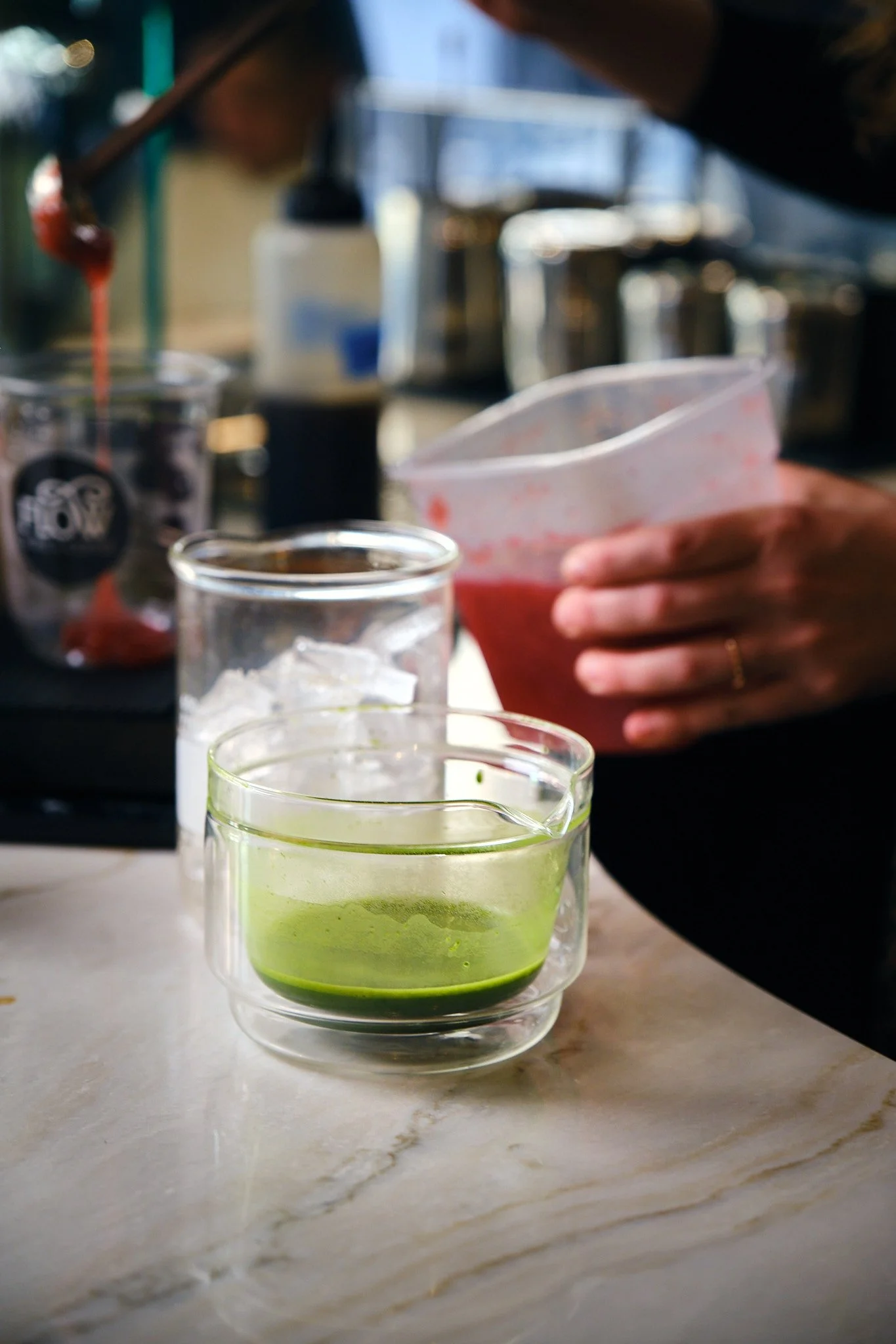 A person holding a plastic container with a pink liquid, surrounded by glasses and bottles on a kitchen counter.