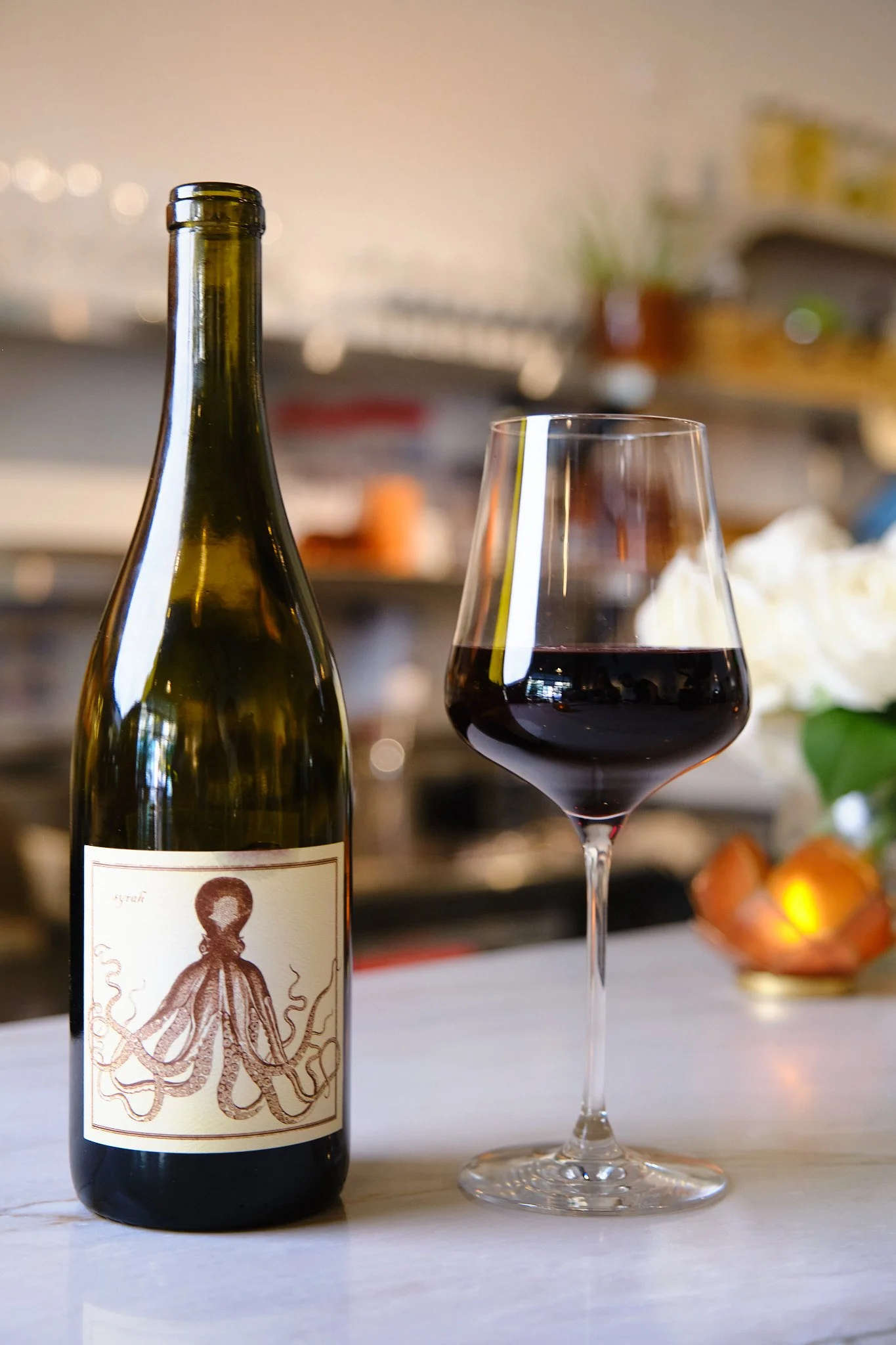 A bottle of wine and a glass of red wine on a white surface with blurred background of dishes and plants.