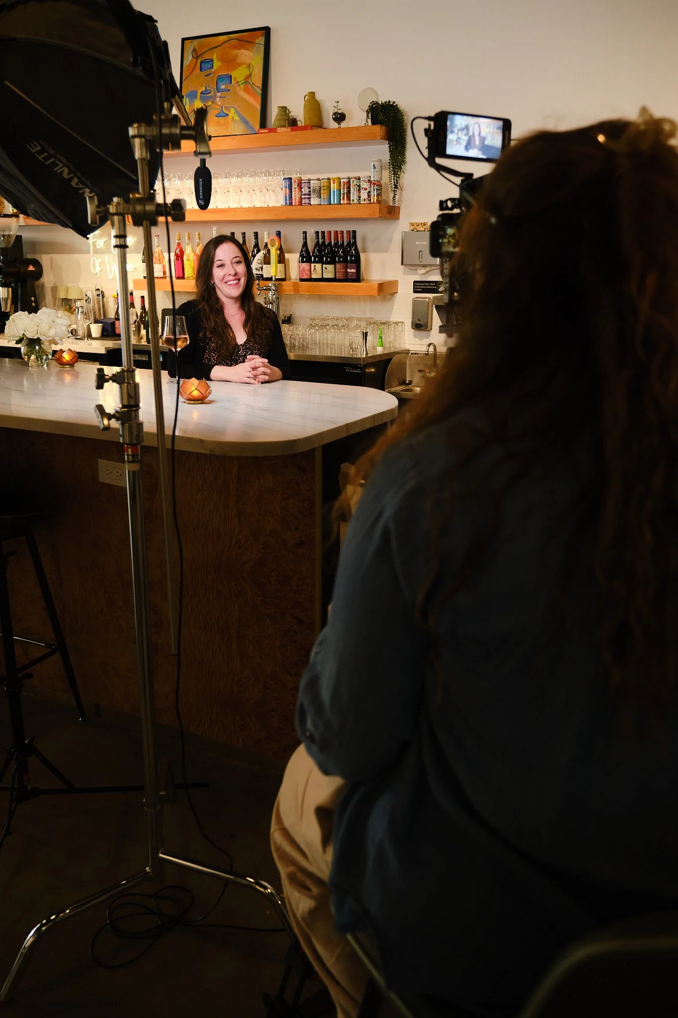 A woman with long brown hair sitting at a white marble bar, smiling and holding a glass of rosé wine. Behind her are shelves with wine bottles, cans, and glassware. The setting appears to be a cozy, modern bar or restaurant with artwork on the wall a