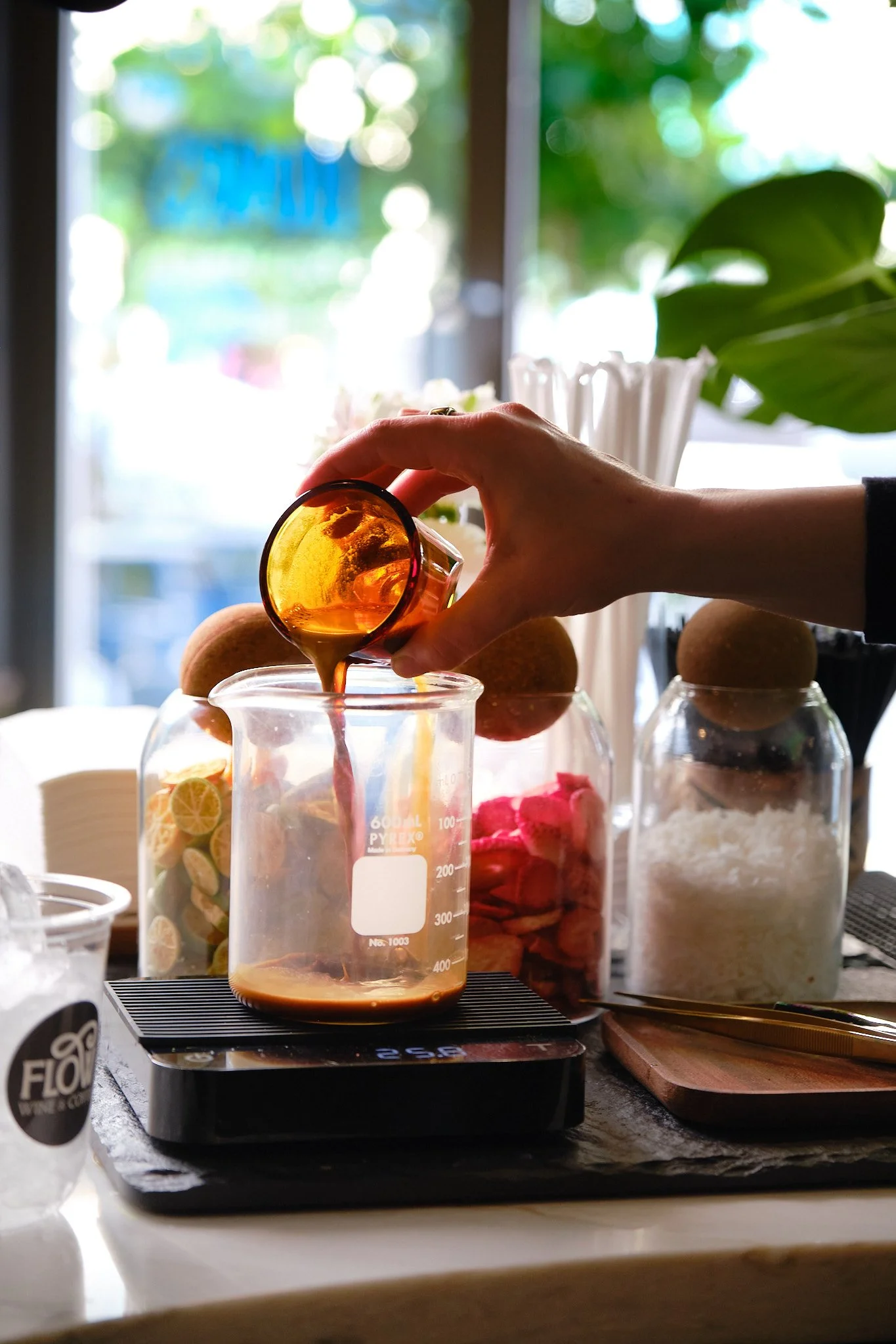 Pouring a caramel sauce from a small glass into a measuring jar on a black stove, with jars of ingredients and a plant in the background.