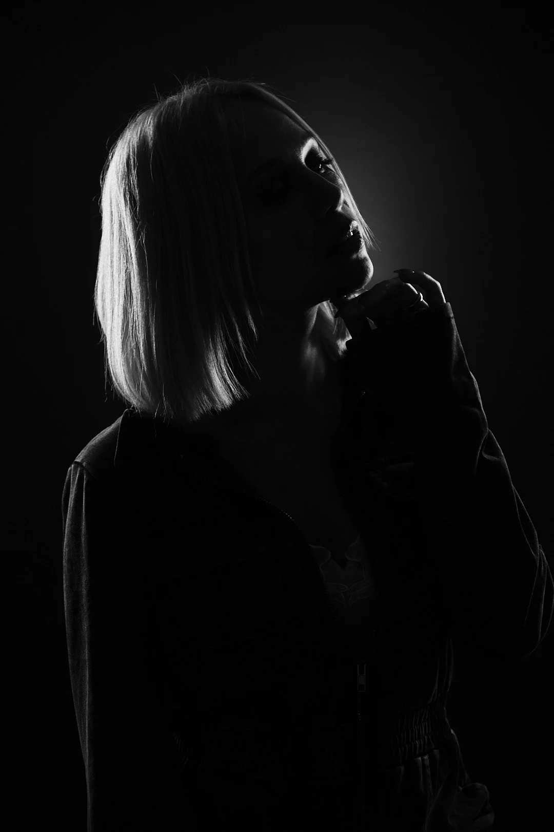 Black and white silhouette of a woman with shoulder-length hair, wearing a jacket, touching her chin with her hand, against a dark background.