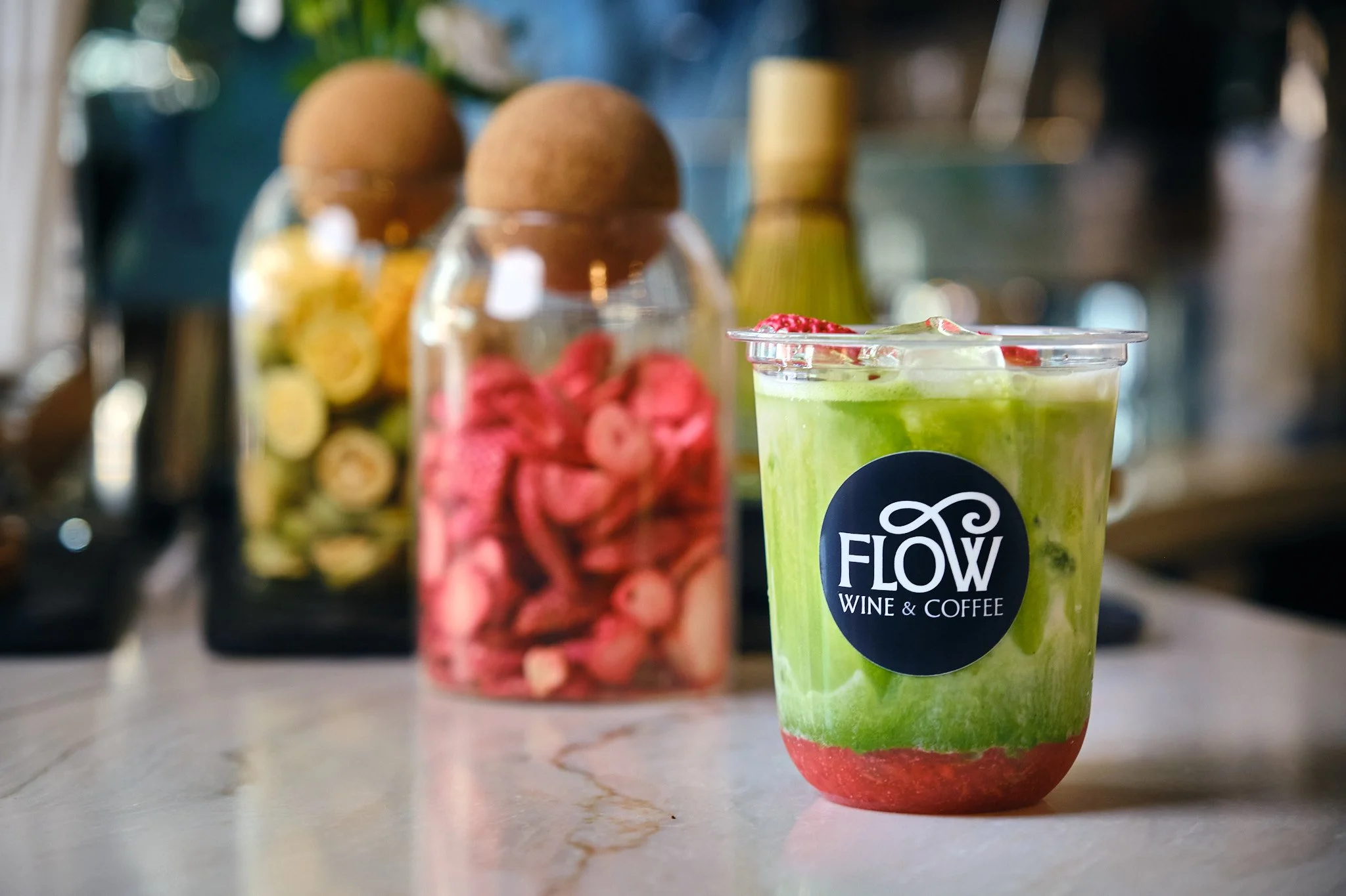 A green smoothie in a clear cup with a black circular sticker that reads 'FLOW WINE & COFFEE' positioned on a marble countertop. In the background, there are bottles, one with the appearance of eggs and others with colorful contents, slightly out of 