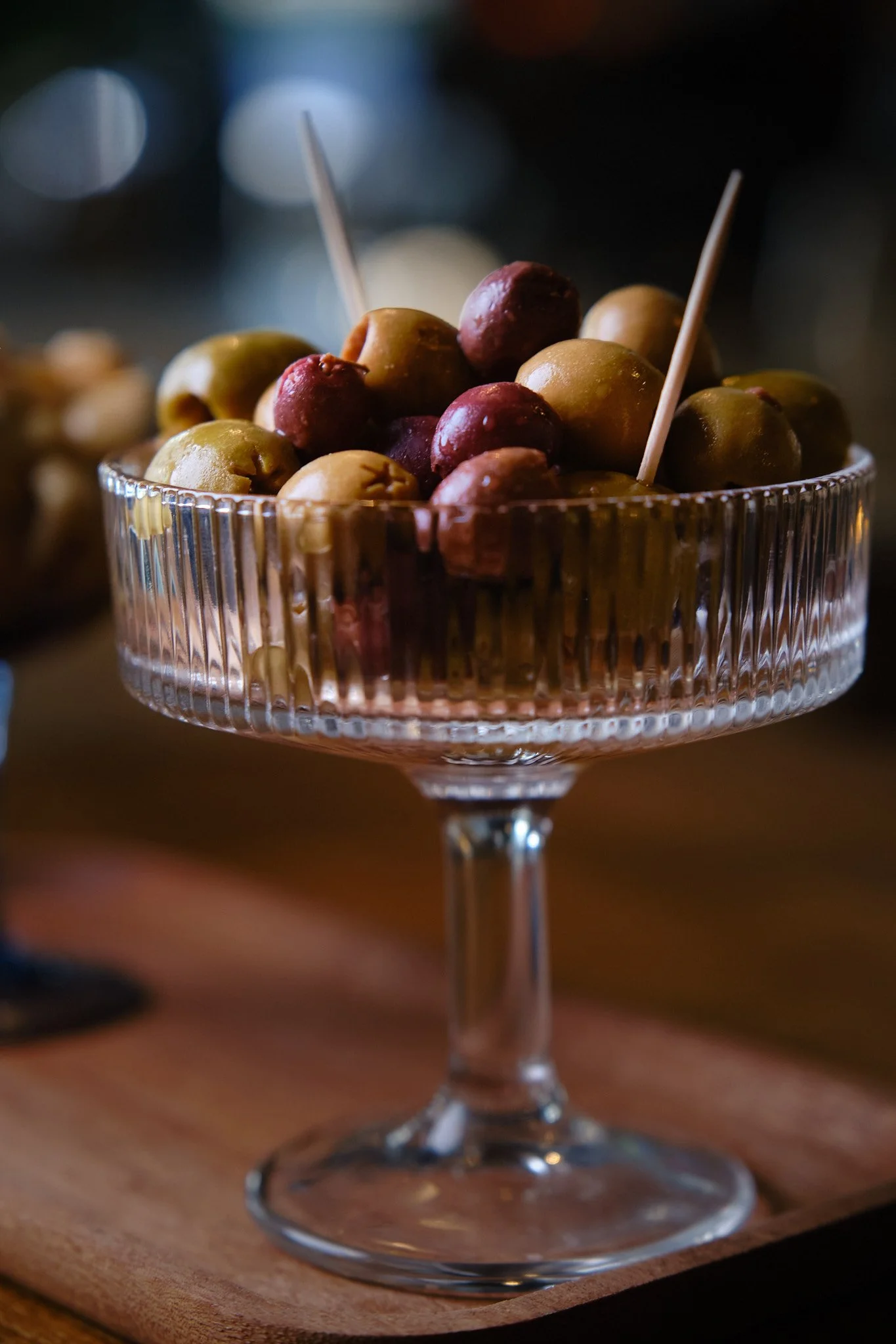A crystal dessert bowl filled with green and purple olives, placed on a wooden surface.