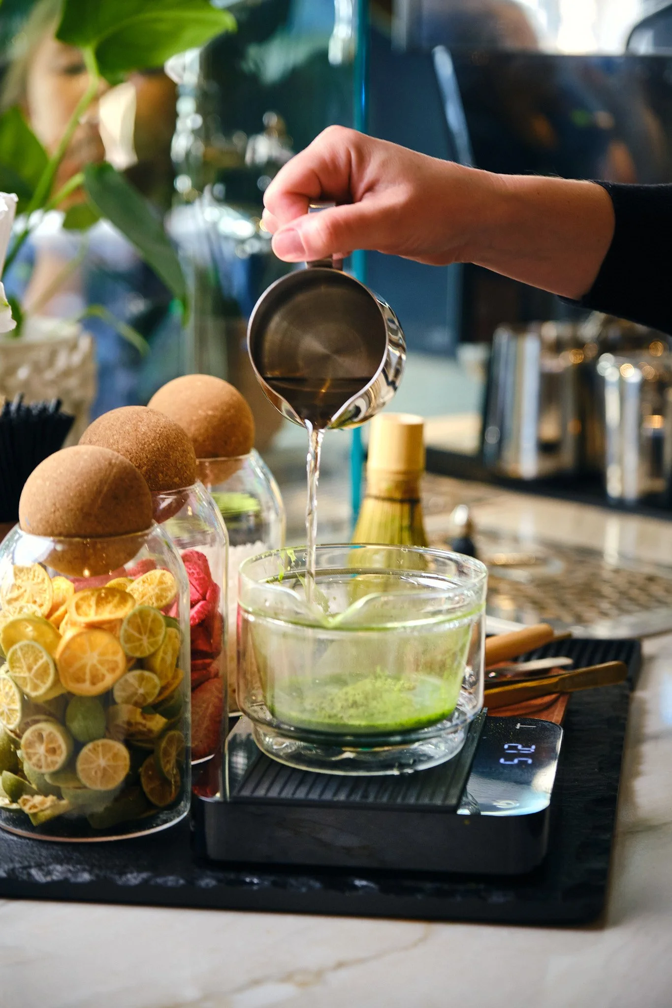 A person pouring a liquid into a glass on a digital scale. There are jars of dried lemon slices and red dried fruit, and a glass of green beverage with lime slices on a table.