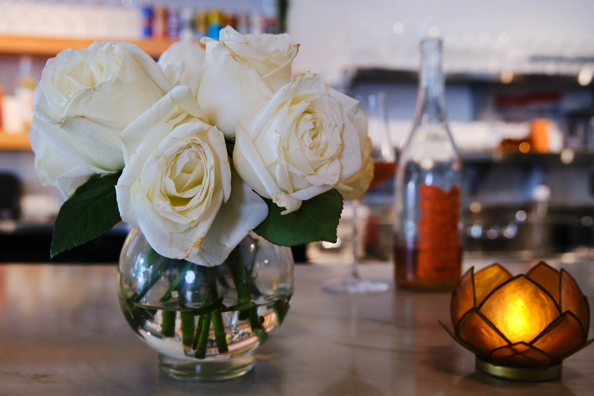 A glass jar filled with white roses on a table, with shelves and bottles in the background, and a glowing orange candle holder to the side.