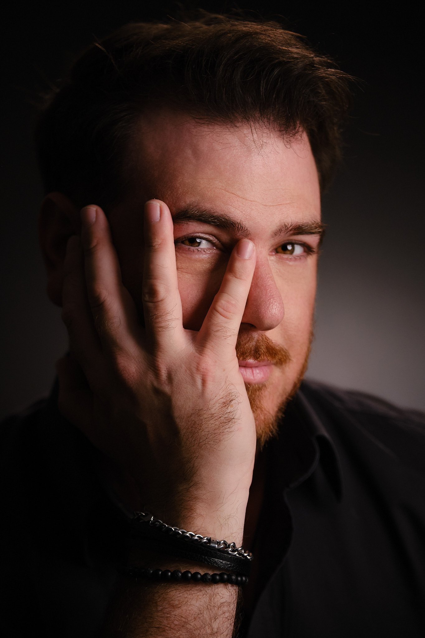 A man with short brown hair and a beard, partially covering one eye with his fingers, looking directly at the camera. He is wearing a black shirt and a chain necklace, set against a dark background.