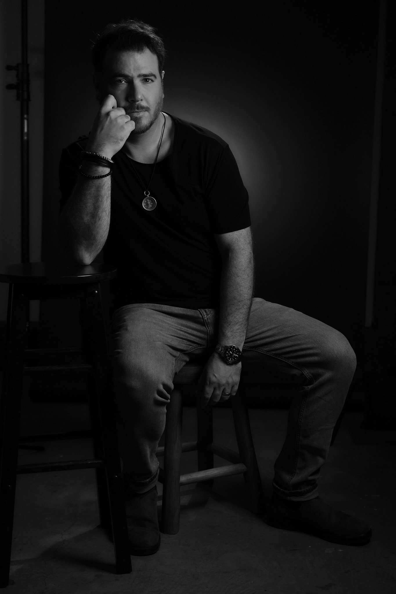 A man with a beard and short hair wearing a black t-shirt, sitting on a wooden chair, looking at the camera, with one hand resting on his face and wearing a watch. The background is dark and the photo is in black and white.
