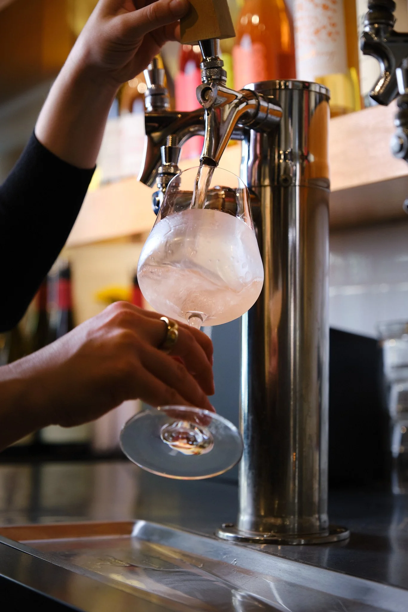 A person pours water into a wine glass from a chrome tap in a bar or restaurant setting. Bottles are visible in the background.