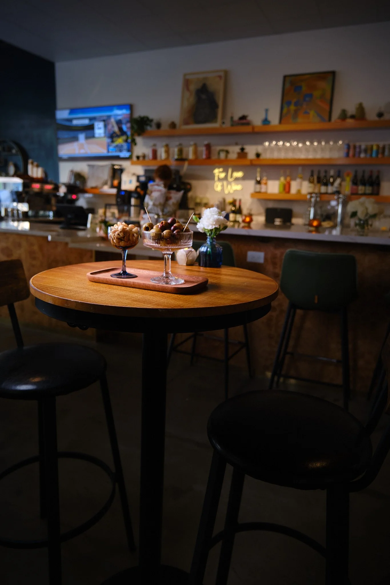 A cozy cafe or bar with a wooden round table in the foreground holding two glasses of snacks, with a white flower in a blue vase. The background features shelves with bottles, glasses, and artwork, a TV screen, and a person behind the counter prepari
