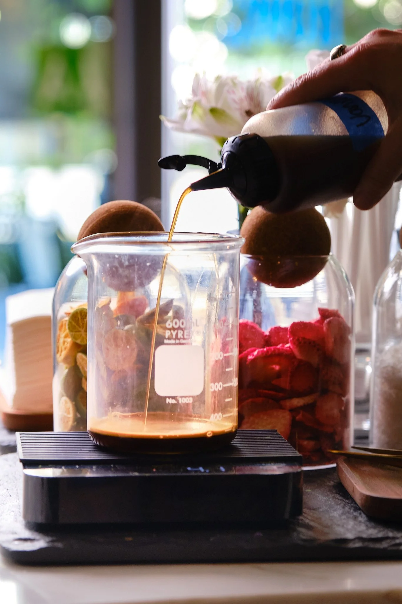 A hand pouring syrup into a glass pitcher with fruit chips inside, with jars of cookies and flowers in the background.