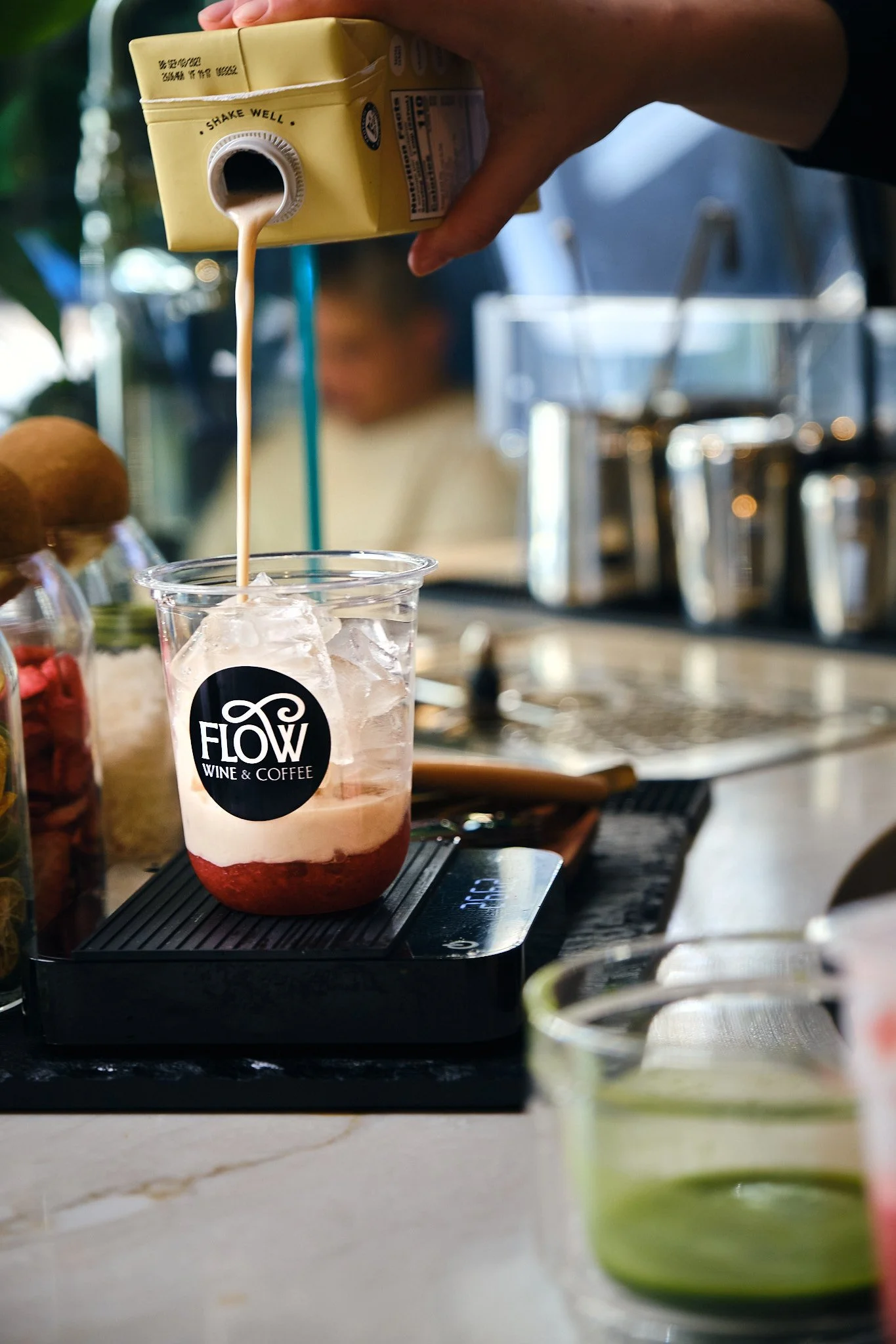 A person pouring a creamy beverage from a yellow carton into a glass with ice, on a black tray in a cafe setting.