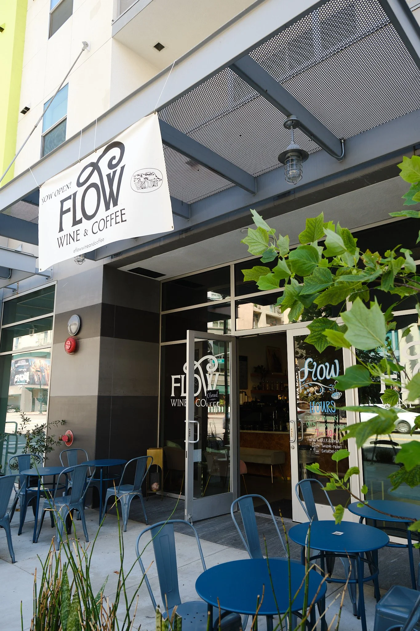 Outside of a cafe named Flow Wine & Coffee with seating area, blue metal chairs and tables, green plants in the foreground, and a glass door entrance.