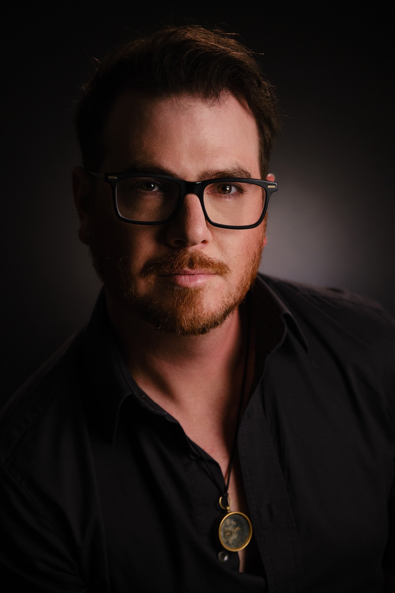 A close-up portrait of a man with brown hair, a beard, and glasses, wearing a black shirt, against a dark background.