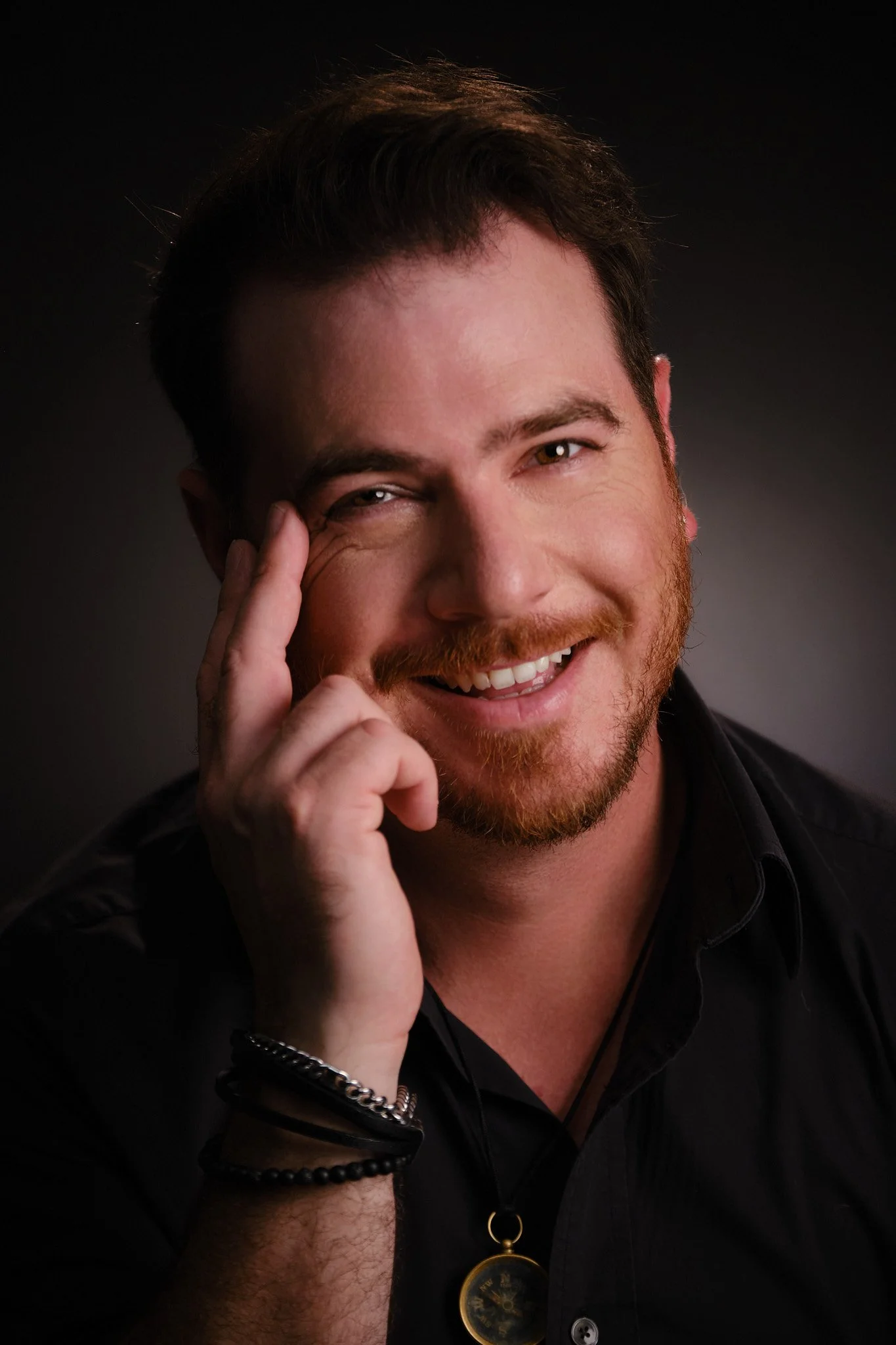 A man with a beard and mustache smiling while touching his forehead, wearing a black shirt, black bracelets, and a pocket watch hanging from his pocket.