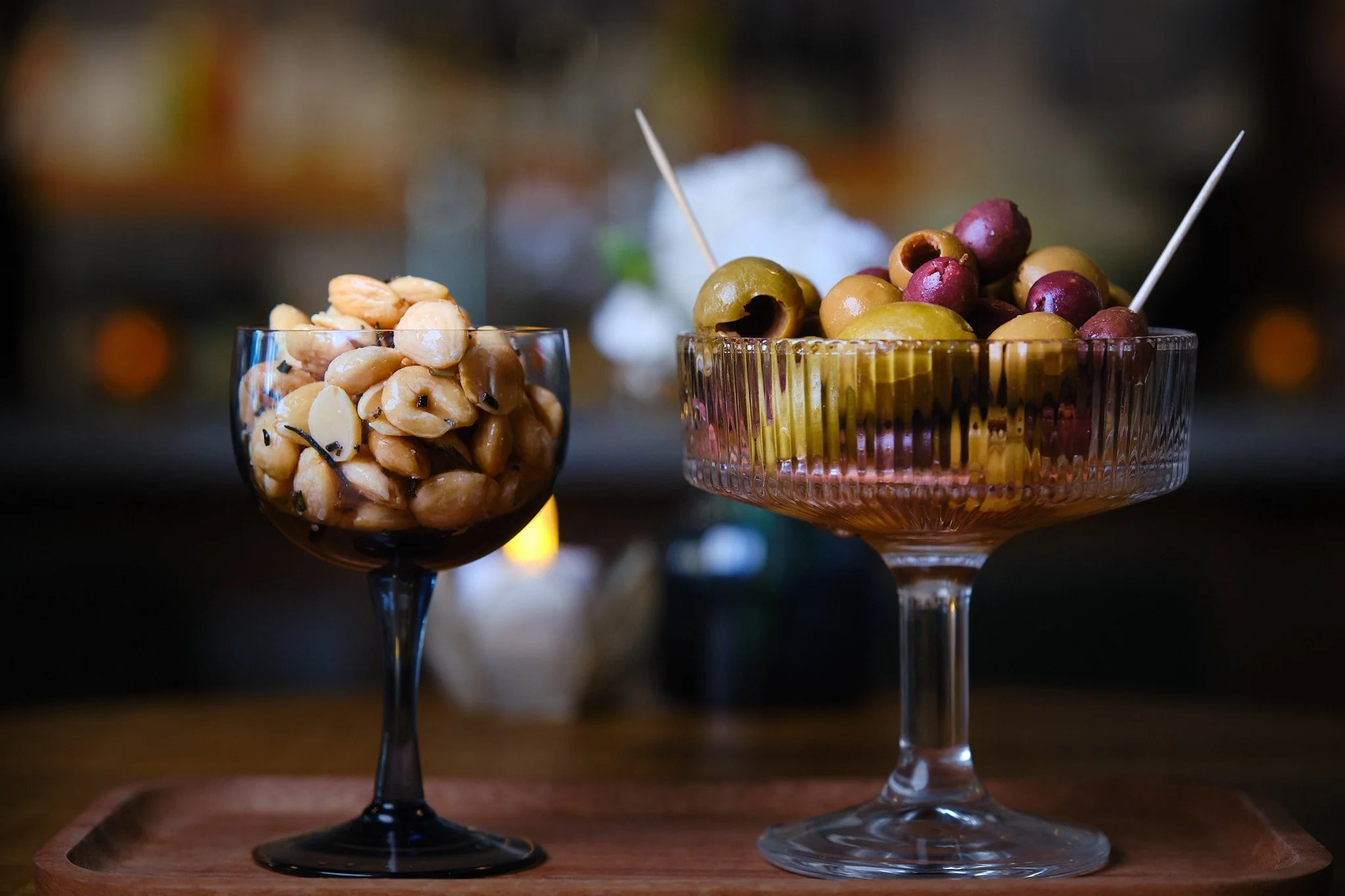 Two glass bowls filled with assorted pickled olives and marinated nuts, set on a wooden tray with a dark blurred background.