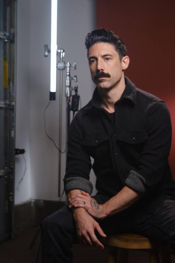 A man with dark hair, a mustache, and tattoos on his left hand, wearing a black jacket, sitting on a wooden stool in front of a red wall with studio lighting equipment in the background.