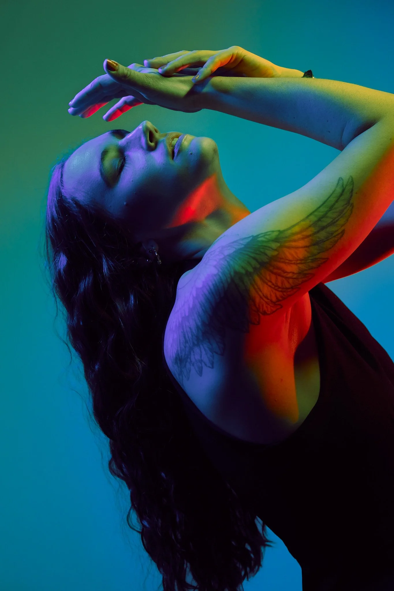A woman with long, curly hair closed her eyes and tilted her head back. She has tattoos of wings on her upper arm and is making a pose with her hands near her face. The image features colored lighting with shades of green, blue, purple, and red, crea