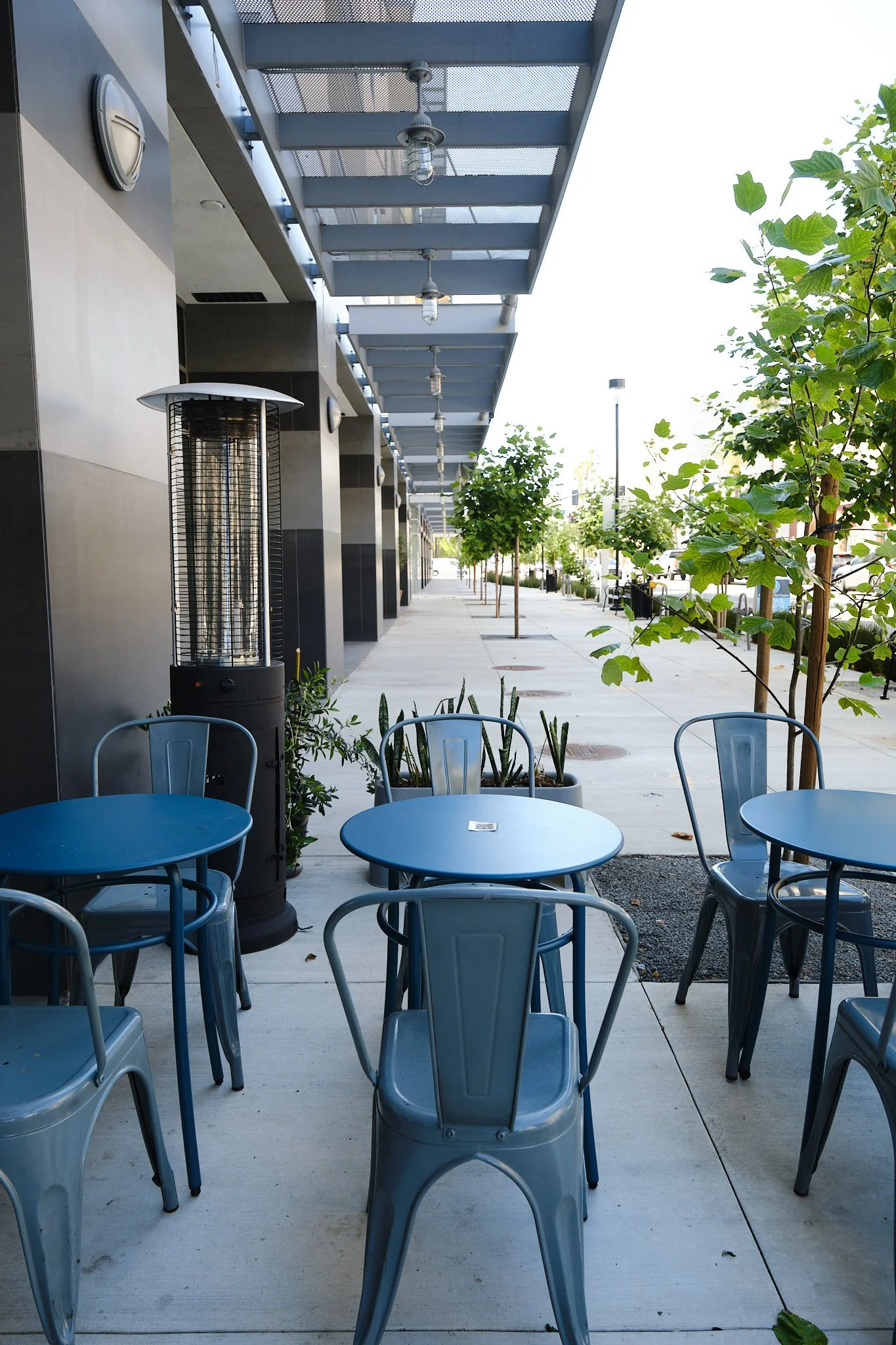 Empty outdoor seating area of a cafe or restaurant with blue chairs and tables, green trees lining the sidewalk, and a patio heater in the foreground.