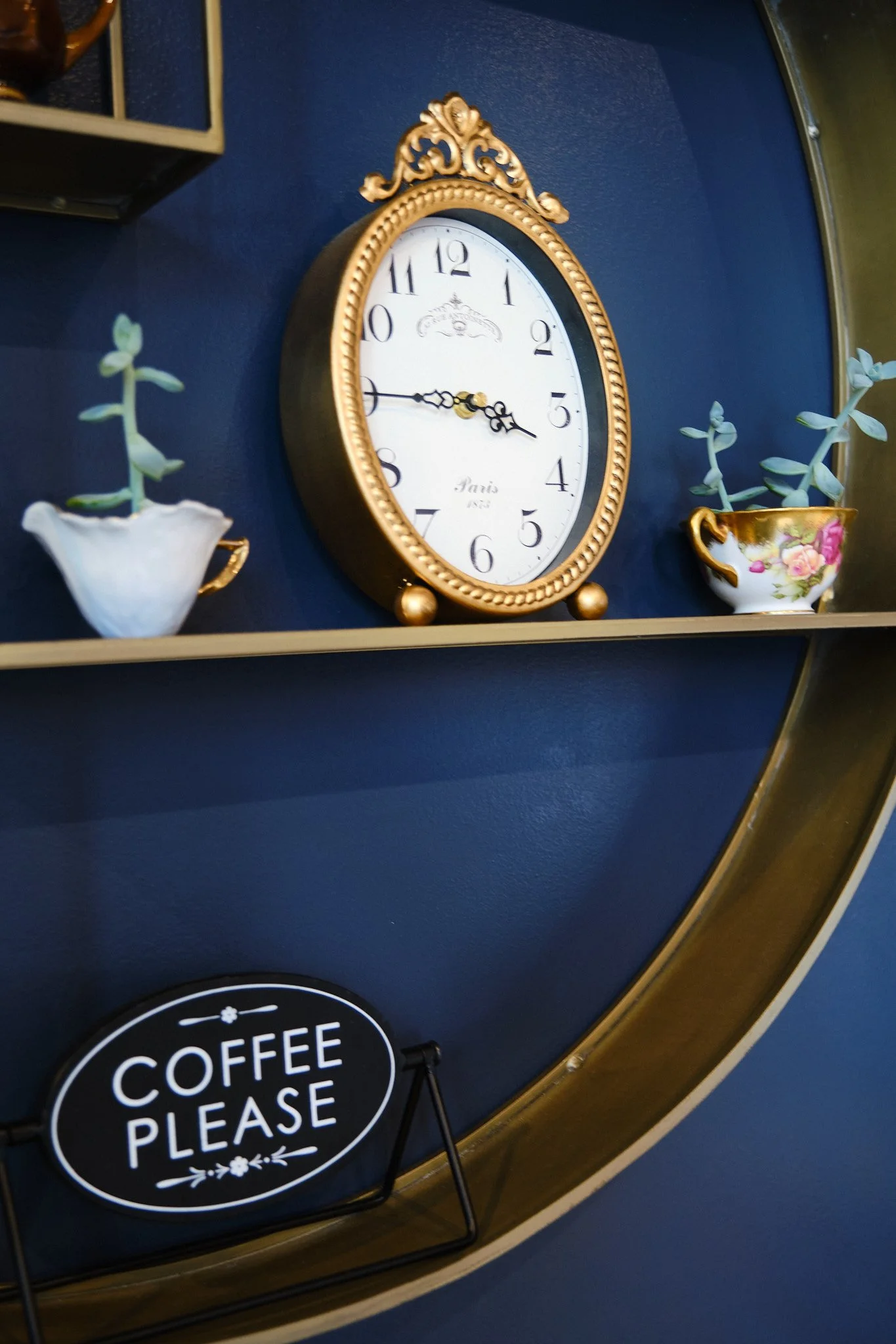 Decorative gold-framed clock on a blue wall with two flower vases, one with succulent plant and the other with pink flowers, and a sign that says 'COFFEE PLEASE'.