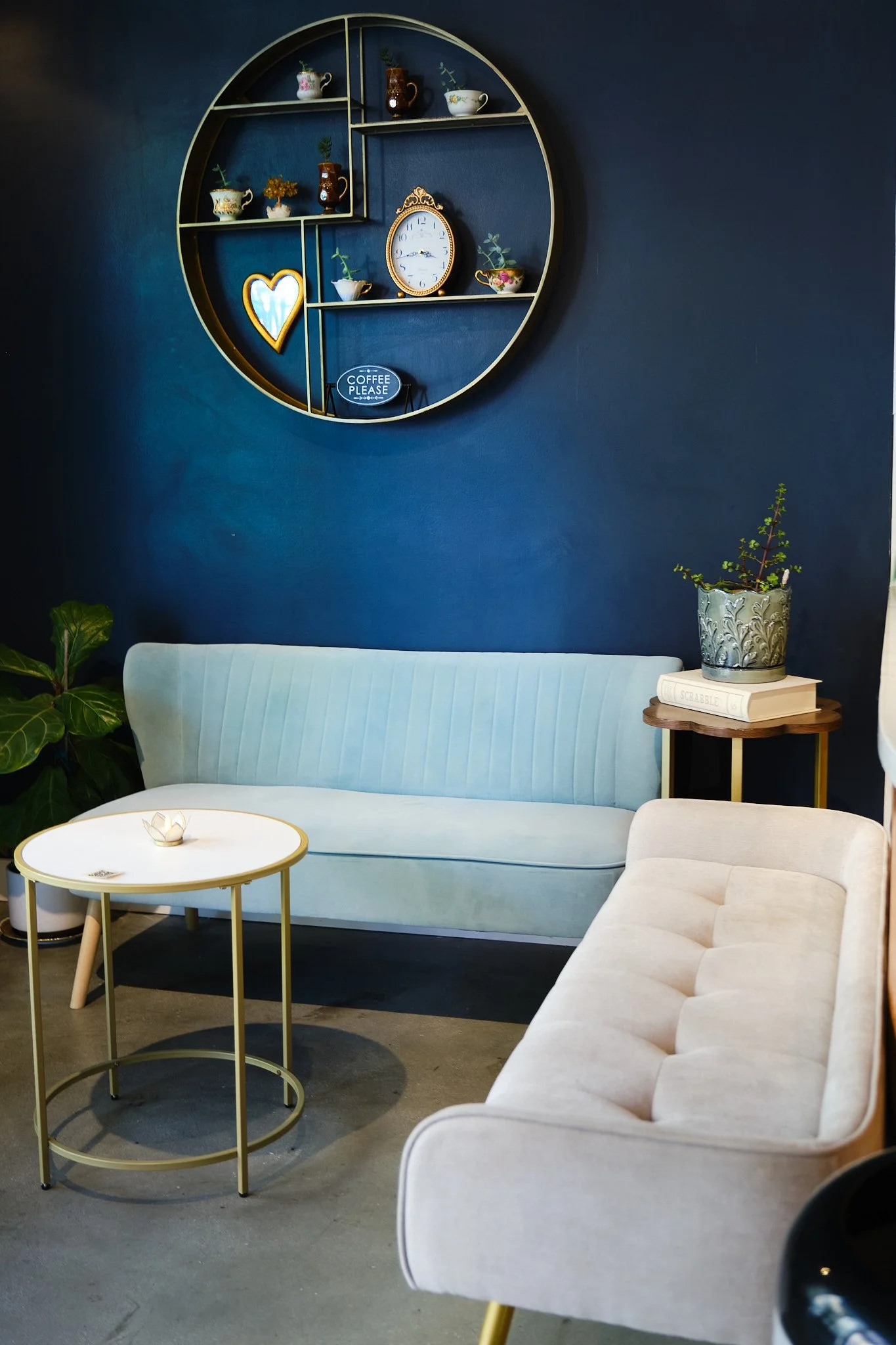 Interior of a modern living room with a blue wall, a white curved sofa, a round coffee table with a small bowl, potted green plants, a bookshelf with decorative items, including small vases, cups, a clock, and a neon heart sign, creating a cozy atmos