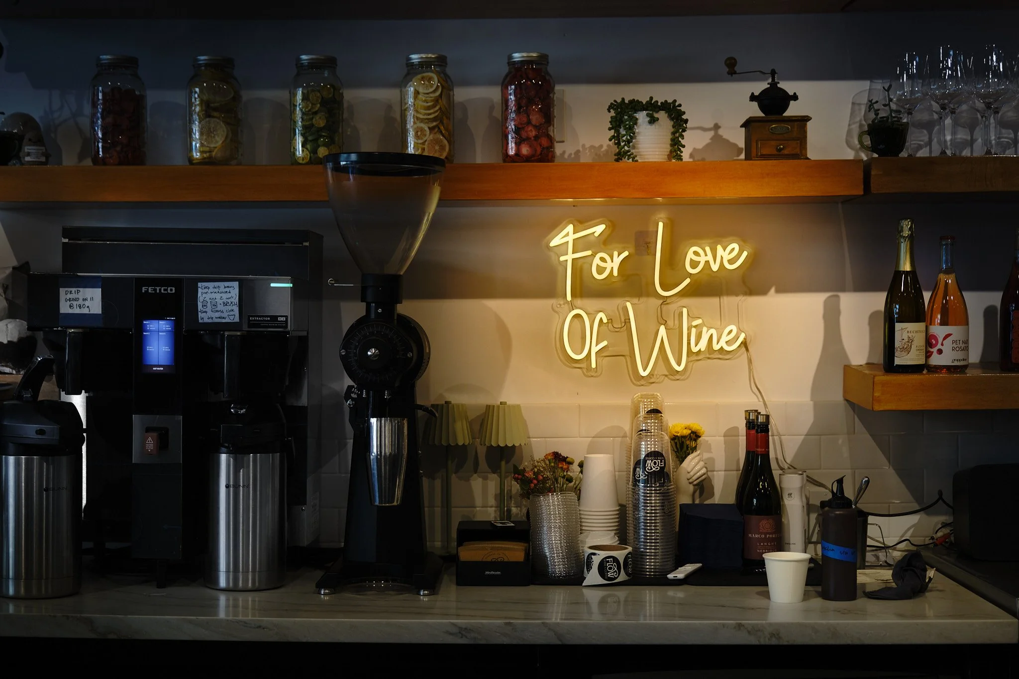 Barista station with a coffee grinder, coffee cups, and bottles of wine, decorated with a neon sign that says 'For Love of Wine' and jars of preserved lemons and other fruits on a shelf above.