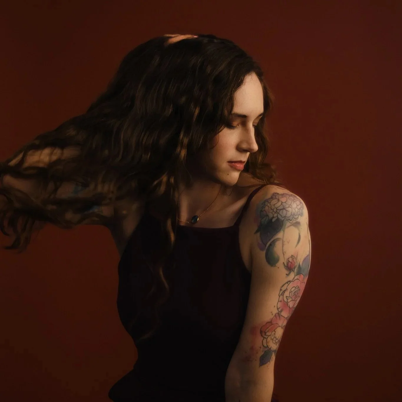 A woman with long, wavy brown hair looking down and to the side. She is wearing a black sleeveless top and has colorful floral tattoos on her upper arm. The background is a solid warm brown color.